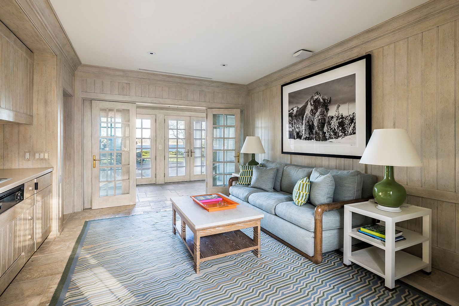 This living room features a cozy and inviting atmosphere with a light blue sofa, a patterned rug, and a coffee table with a marble top. Natural light floods the space through the French doors, and a large black and white photograph hangs above the sofa. Two green lamps sit on either side of the sofa, adding a pop of color.