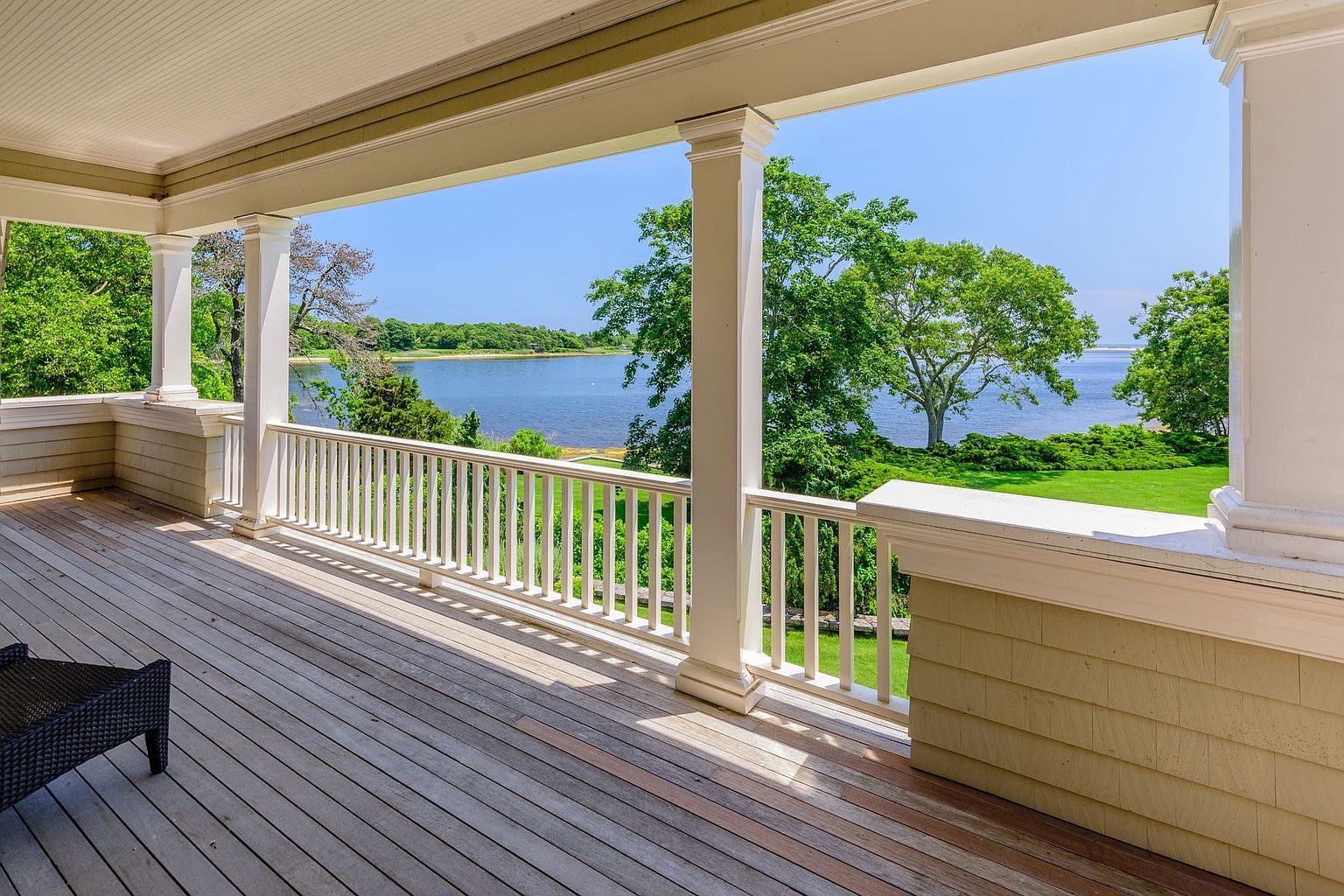 This image showcases a spacious outdoor patio or deck area with a stunning waterfront view. The deck features wooden flooring and a white railing, complemented by white support columns. The scene offers a serene and luxurious ambiance, perfect for relaxation and enjoying the natural surroundings.