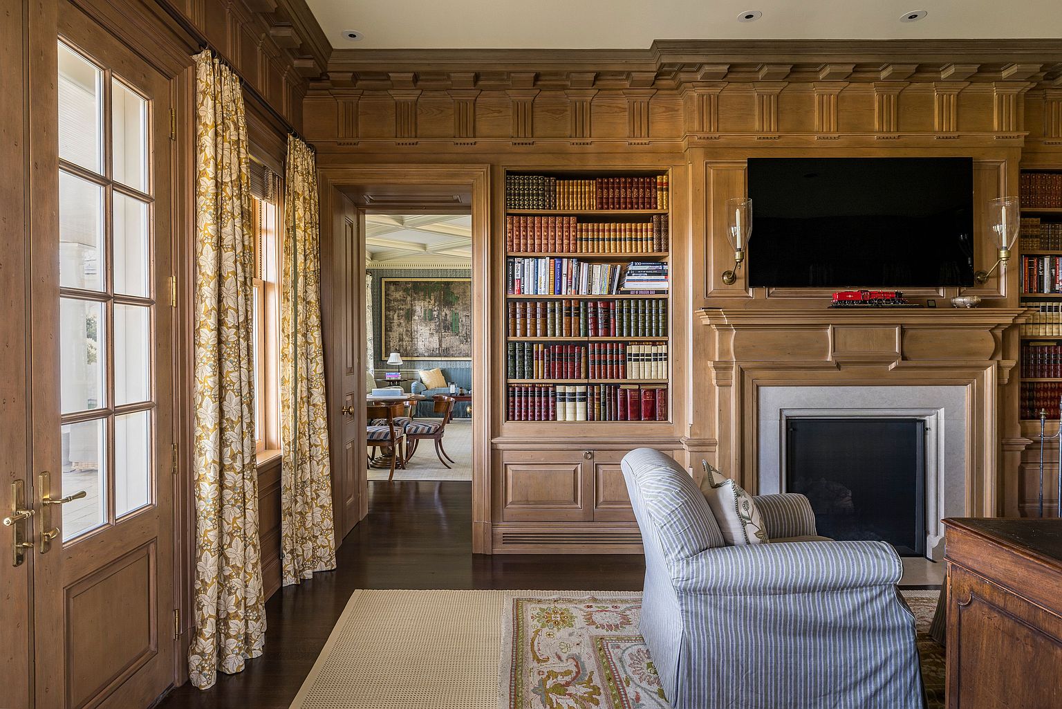 This is an interior shot of a traditional living room featuring wood paneling, built-in bookshelves, and a fireplace with a television mounted above. A comfortable armchair sits on an area rug, and a doorway leads to another room, creating a sense of depth and spaciousness. The room exudes a warm and inviting atmosphere, perfect for relaxation and entertainment.