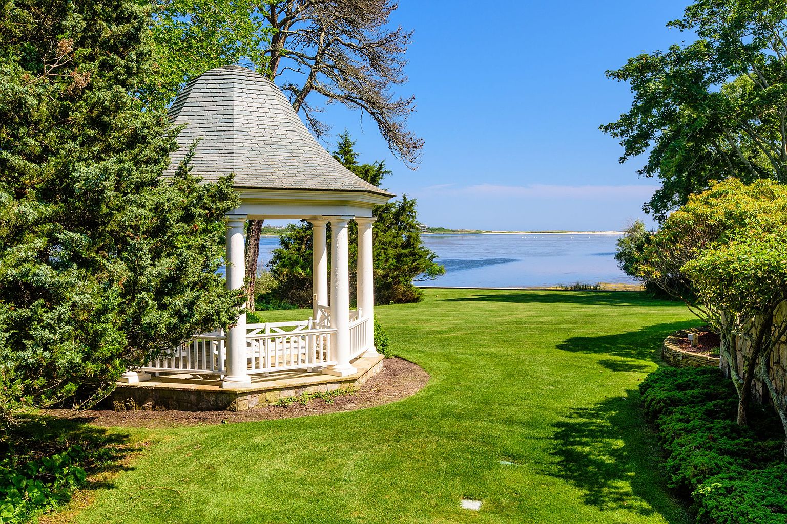 This image showcases a beautifully landscaped yard featuring a charming gazebo as a focal point. The lush green lawn extends towards a serene body of water, creating a tranquil and picturesque setting. Mature trees and manicured shrubs add to the property's appeal, suggesting a well-maintained and inviting outdoor space.