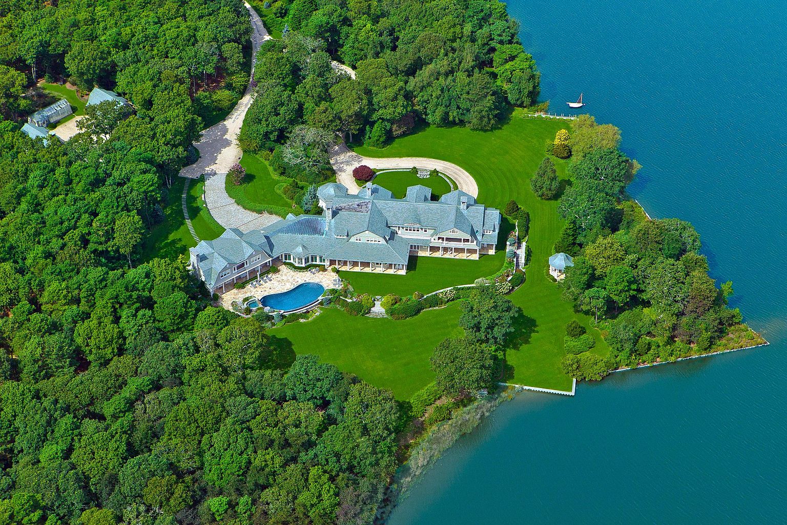 This aerial view showcases a luxurious waterfront estate with a sprawling mansion, meticulously manicured lawns, and a private pool. The property is surrounded by lush greenery and offers stunning water views, creating a serene and exclusive atmosphere. A long driveway leads to the house, emphasizing its grandeur and privacy.