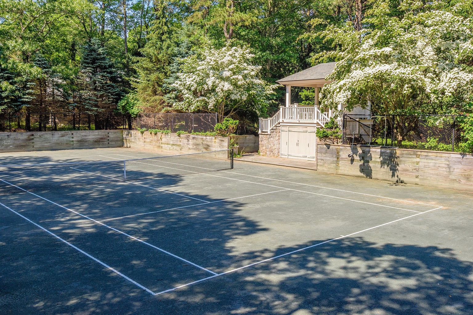 This image showcases a well-maintained tennis court surrounded by lush greenery and mature trees. A charming gazebo-like structure with white railings and stone accents adds a touch of elegance to the landscape. The court itself is in excellent condition, with clear lines and a taut net, suggesting a private and luxurious amenity.