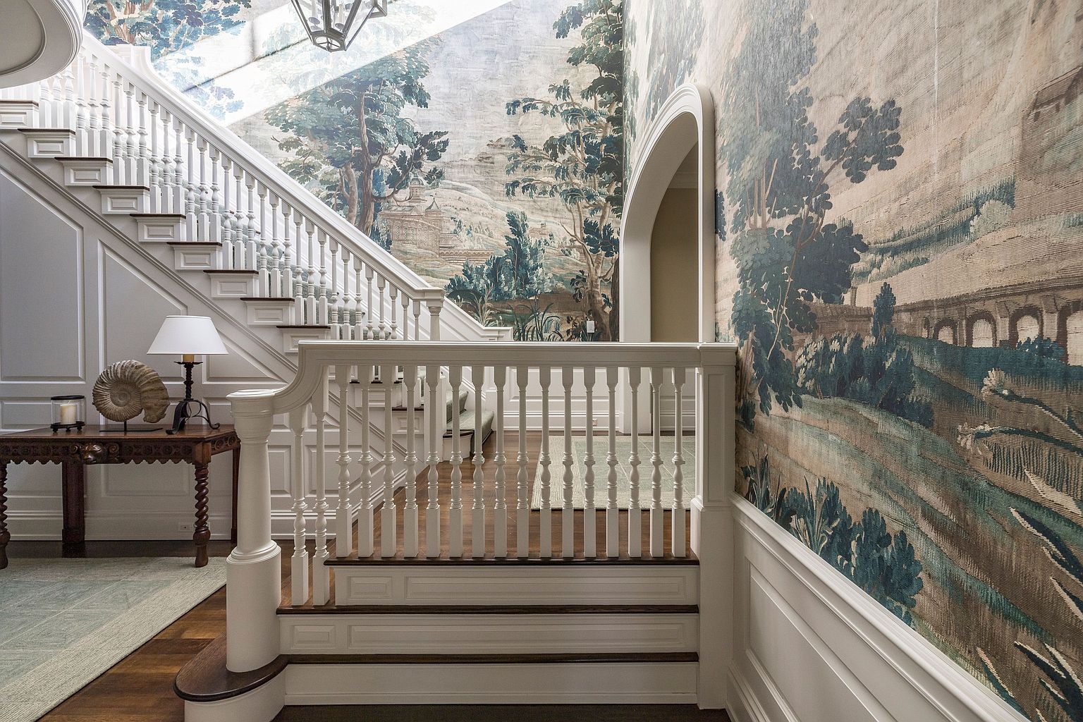 This grand foyer showcases a sweeping staircase with white balusters and dark wood treads, complemented by a scenic mural wallpaper that adds a touch of elegance and sophistication. A decorative table with a lamp and seashell accent piece sits to the side, enhancing the luxurious feel of the space. The overall impression is one of refined taste and timeless design.