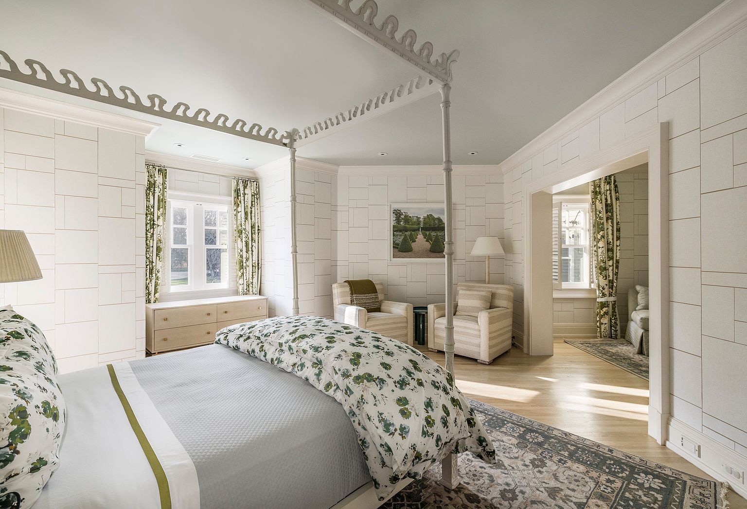This is a bright and airy primary bedroom featuring a four-poster bed with a floral patterned duvet and a gray quilted blanket. The walls are adorned with a unique geometric tile pattern, complemented by matching curtains and a window seat. Two armchairs sit near a landscape painting, creating a cozy seating area.