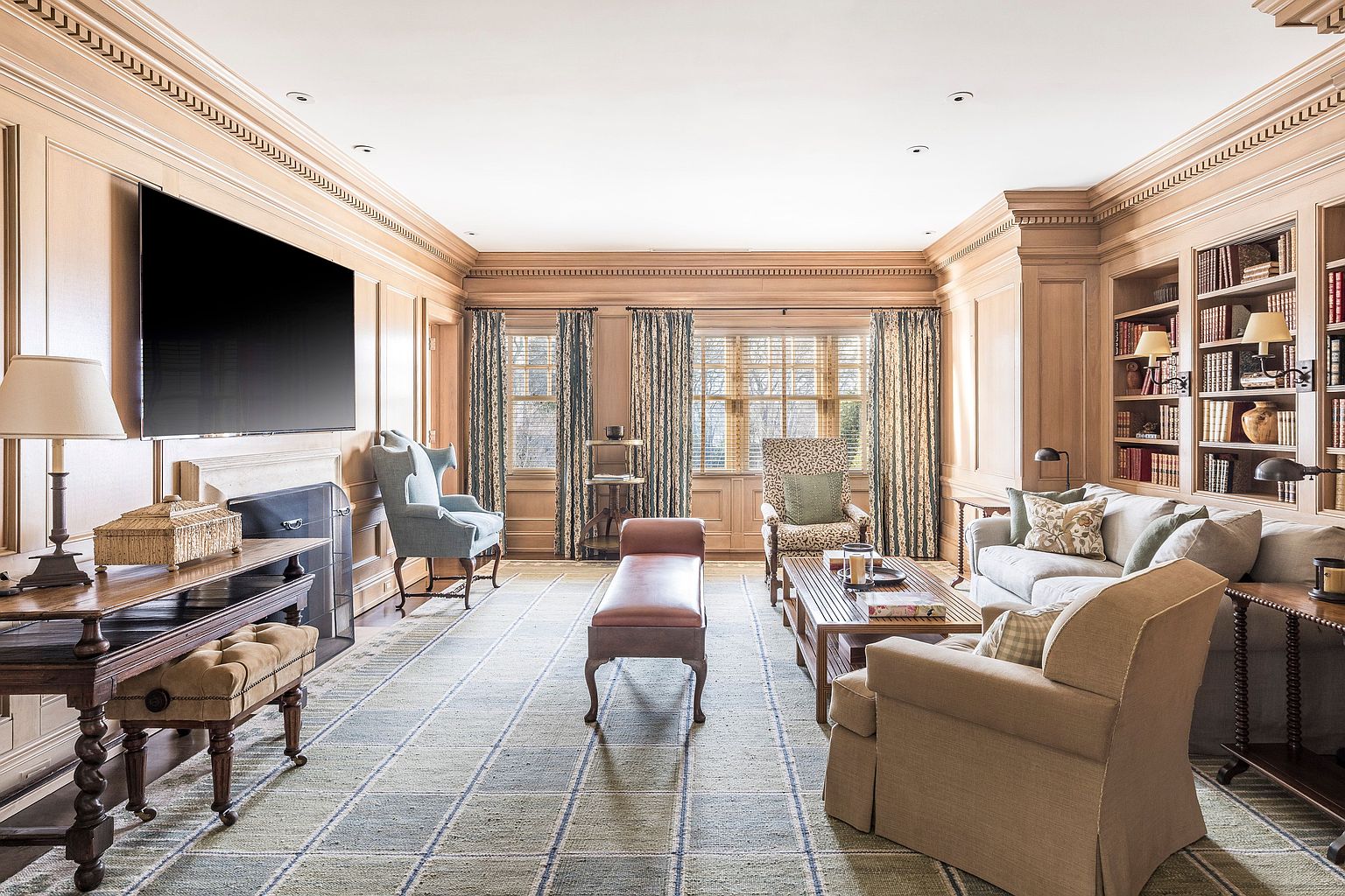 This is a classic living room featuring wood paneling, built-in bookshelves, and a large flat-screen television above a fireplace. The room is furnished with comfortable seating, including sofas, armchairs, and a bench, arranged around a wooden coffee table. A patterned rug covers the floor, and natural light filters through the windows, creating a warm and inviting atmosphere.