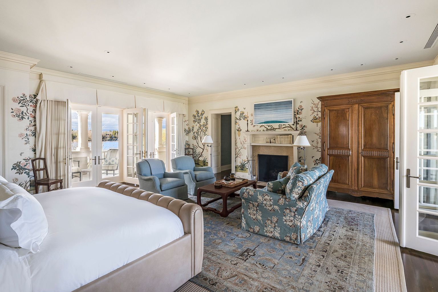 This is a luxurious primary bedroom featuring a king-size bed with a tufted headboard, a sitting area with floral armchairs and a coffee table, and a fireplace with an ornate mantel. The room is decorated with floral wallpaper and has access to a balcony through French doors, offering a serene view. A large wooden wardrobe provides ample storage.