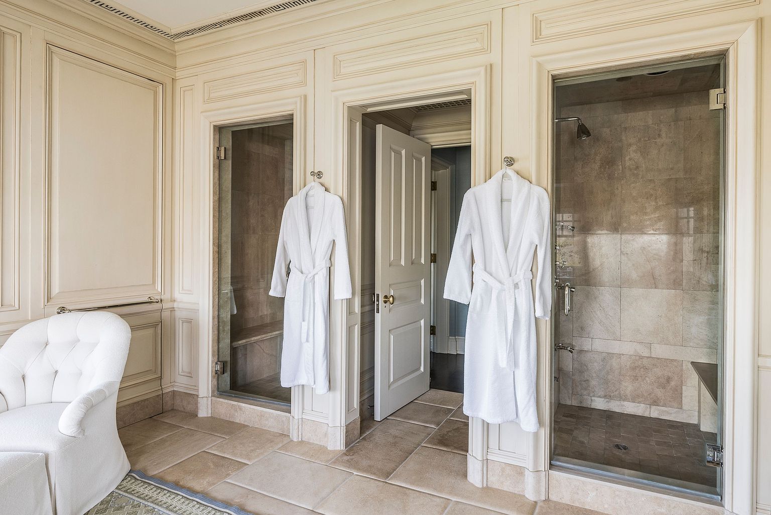 This is a luxurious primary bathroom featuring elegant, paneled walls and tiled flooring. Two glass-enclosed showers flank a central doorway, each with a white robe hanging outside. A white armchair sits to the left, adding a touch of comfort to the sophisticated space.