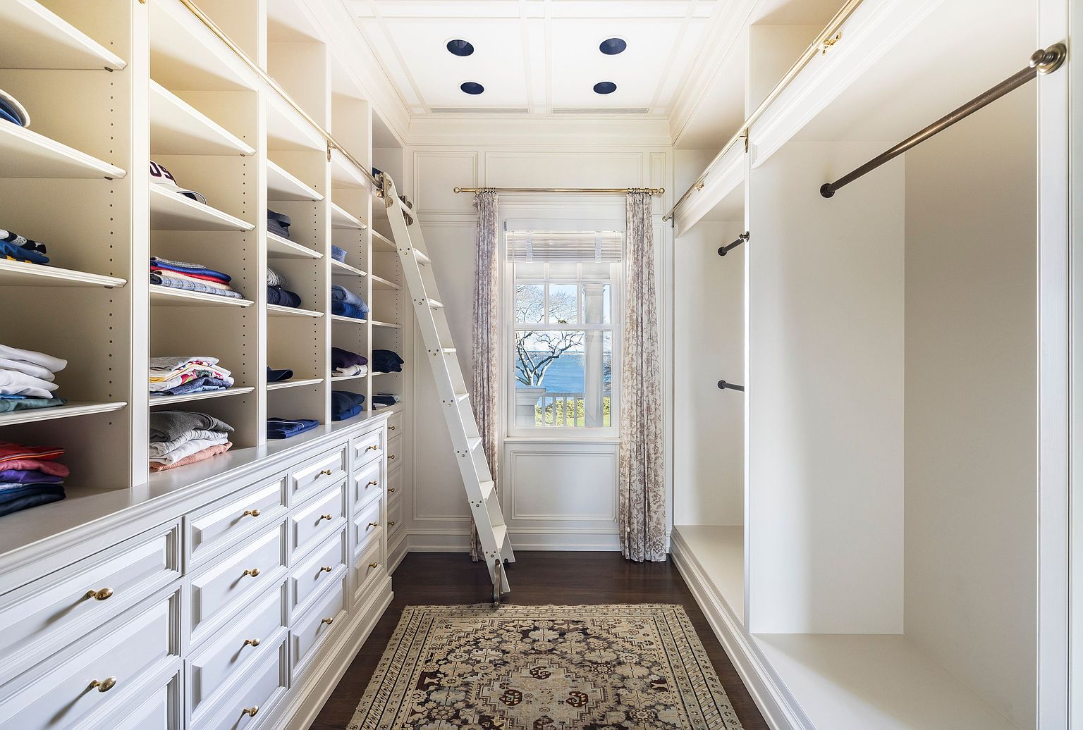 This is a well-organized walk-in closet featuring custom white cabinetry with drawers and shelving. A rolling ladder provides access to higher shelves, and a window offers natural light. The closet also includes hanging rods and a decorative rug, creating a luxurious and functional space.