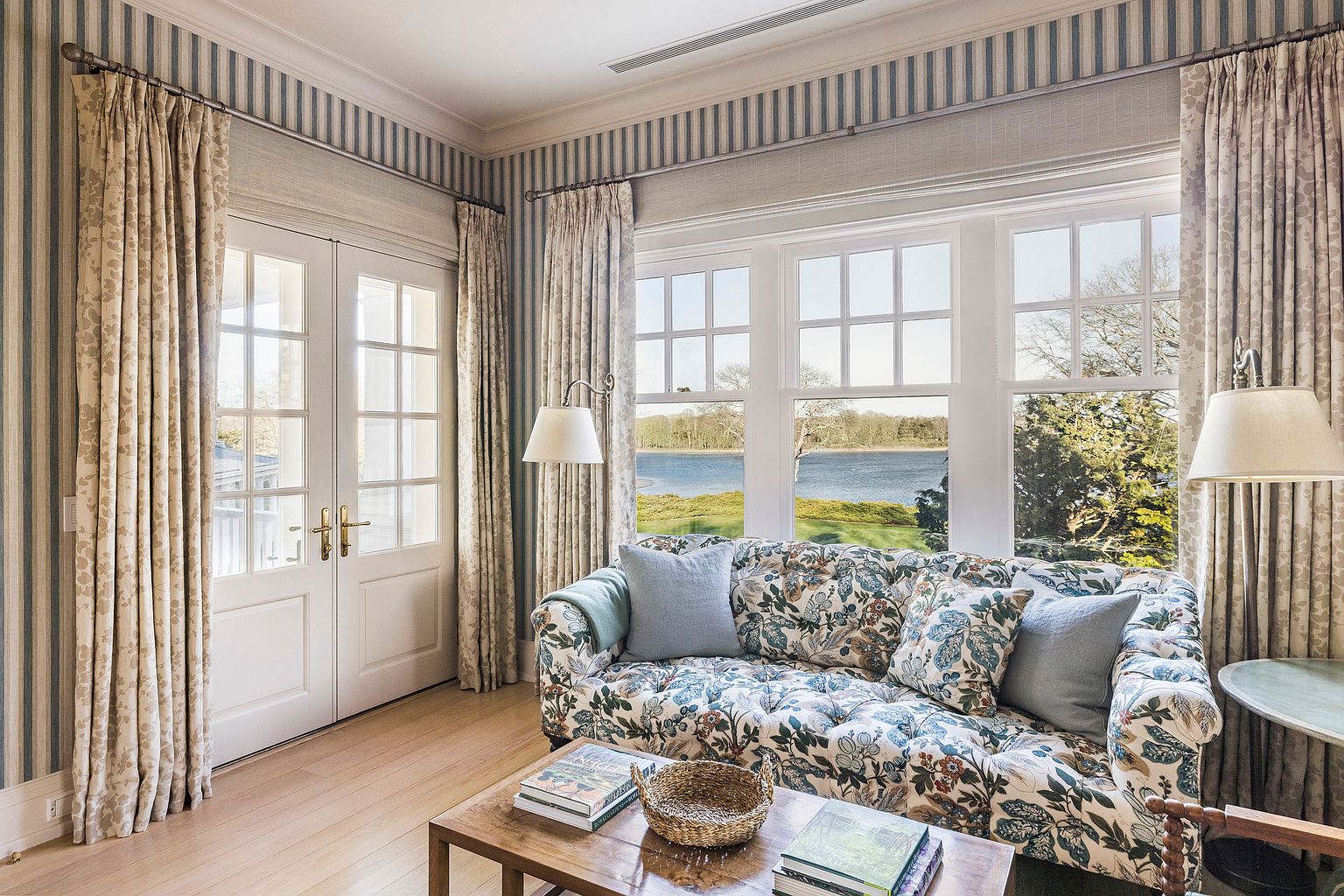 This inviting living room features a floral patterned sofa adorned with light blue pillows, positioned in front of a large window offering a serene view of the water. The room is decorated with striped wallpaper and matching curtains, creating a cohesive and elegant aesthetic. A wooden coffee table sits in the center, adding warmth to the space.