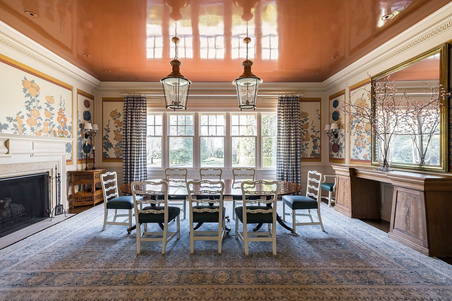 This is a formal dining room featuring a large oval table with seating for eight, set on a patterned area rug. The room is decorated with floral wallpaper panels, plaid curtains, and two hanging lantern-style light fixtures. A fireplace is visible on the left, and a large mirror is on the right, reflecting the room's details and enhancing the sense of space.