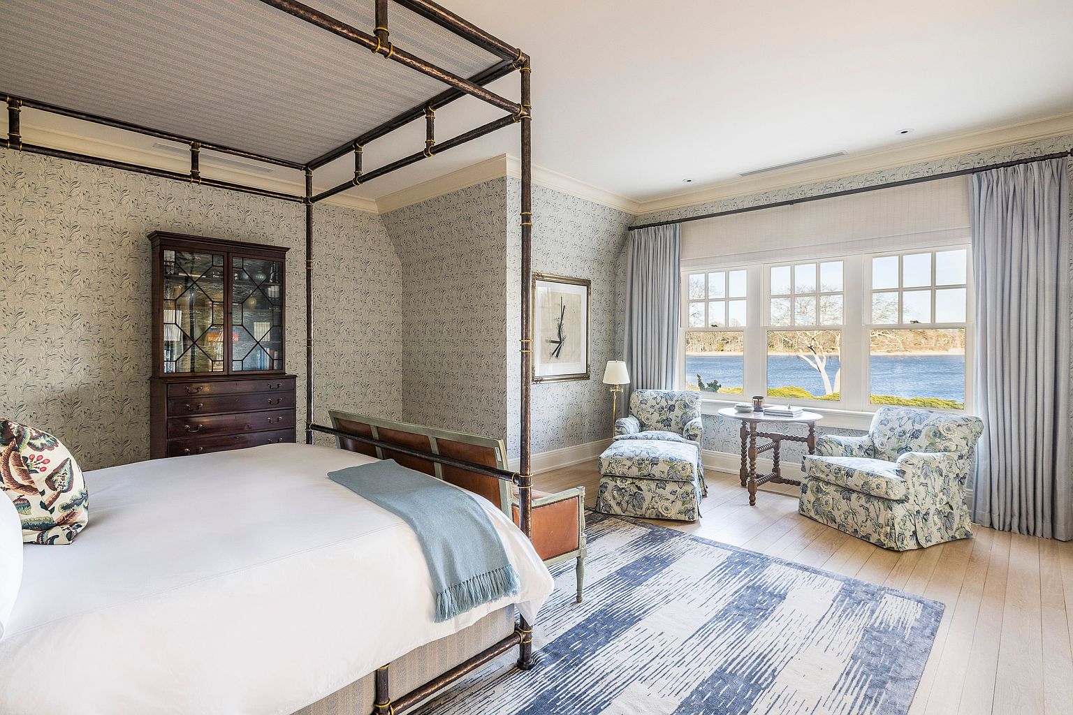 This is a beautifully decorated primary bedroom featuring a four-poster bed with a light blue throw, a dark wood dresser with a glass-front cabinet, and two floral armchairs by a large window overlooking a body of water. The room is decorated with a delicate floral wallpaper and a blue and white striped rug, creating a serene and luxurious atmosphere. The perspective is from the foot of the bed, looking towards the window and seating area.