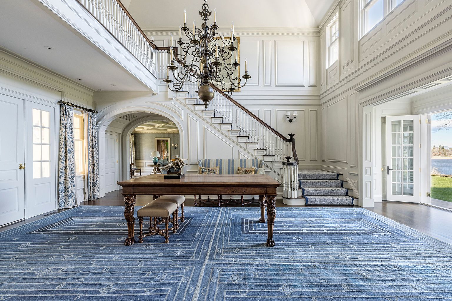 This grand foyer features a soaring two-story entrance with elegant white wainscoting and a striking chandelier. A staircase with a patterned runner leads to the upper level, while a large blue rug anchors the space. The room exudes a sense of luxury and spaciousness, perfect for making a lasting first impression.