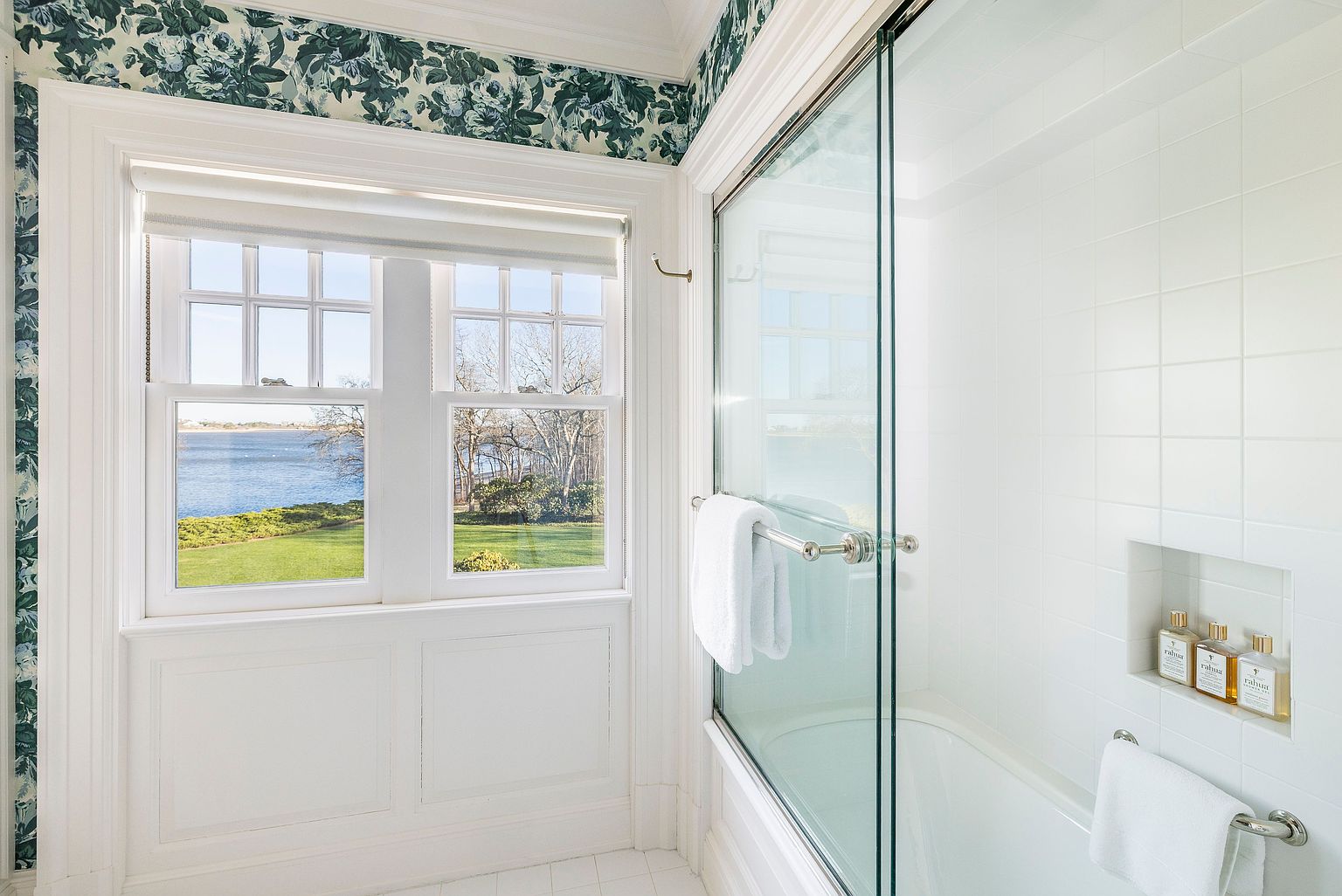 This is a bright and airy primary bathroom featuring a window with a view of the water and greenery. The bathroom has white tile walls, a glass-enclosed shower/tub, and a niche with toiletries. The wallpaper above the wainscoting adds a touch of elegance.