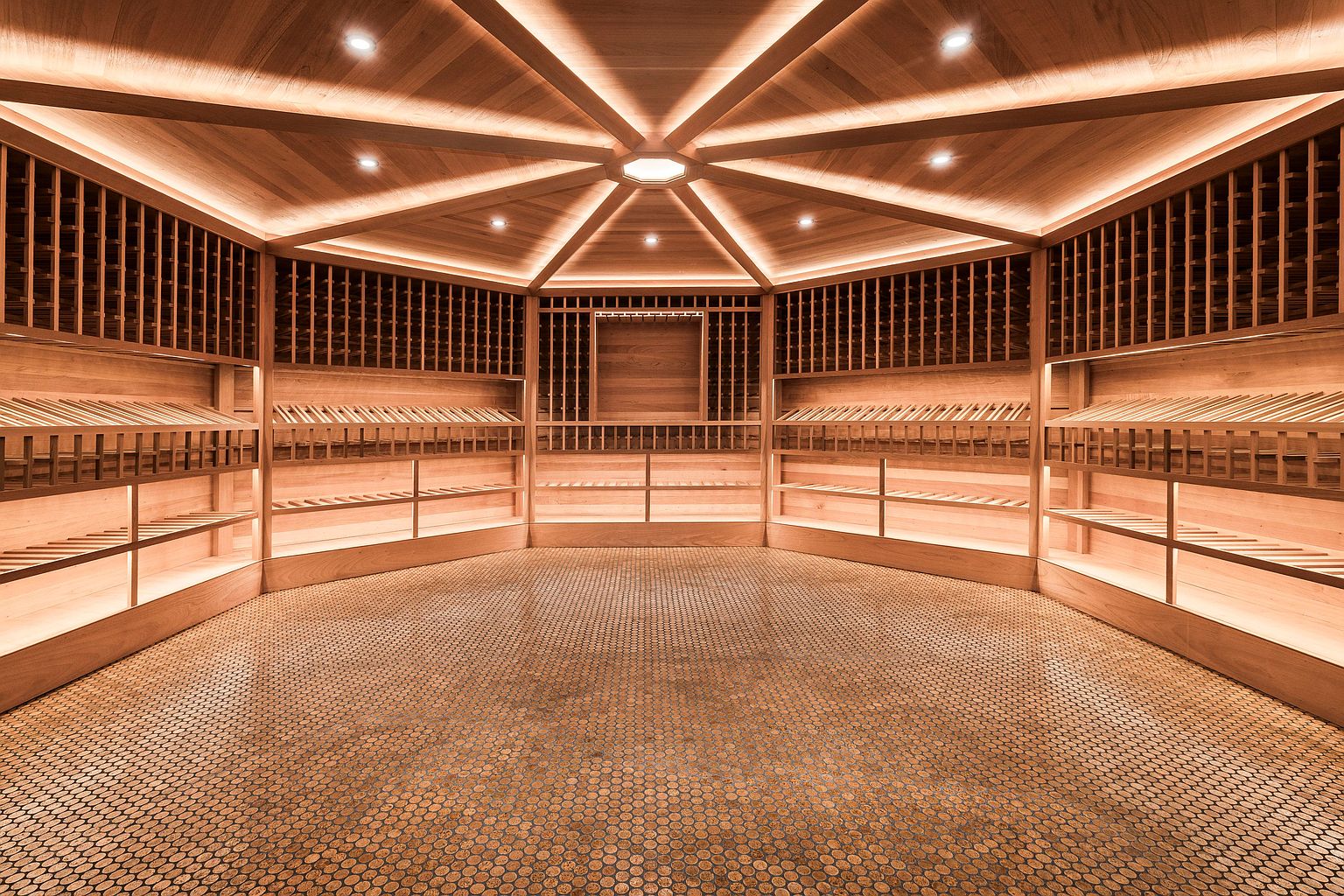 This is an interior shot of a custom wine cellar. The cellar features wooden shelving designed to hold wine bottles, with additional shelving below. The ceiling is a unique, multi-faceted design with recessed lighting, and the floor is covered in small, round tiles, creating a warm and inviting atmosphere.