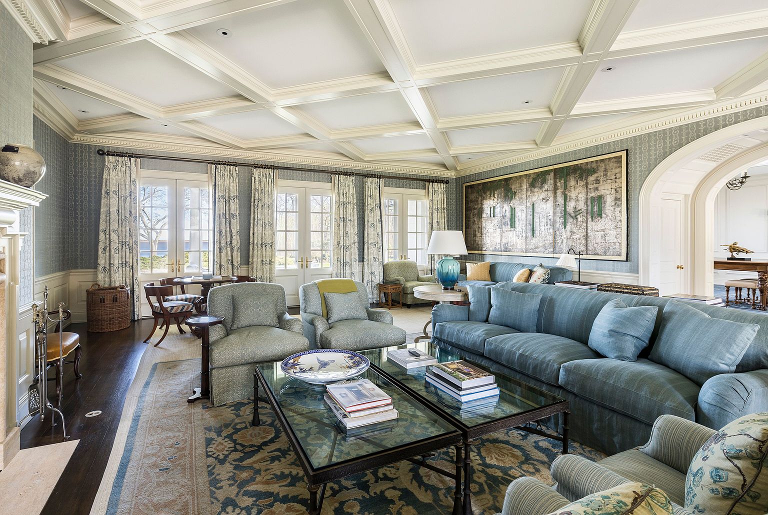 This is a luxurious living room featuring a coffered ceiling, patterned wallpaper, and a large abstract painting. The room is furnished with a blue sofa, patterned armchairs, and a glass-topped coffee table adorned with books. Natural light floods the space through multiple windows, creating a bright and inviting atmosphere.