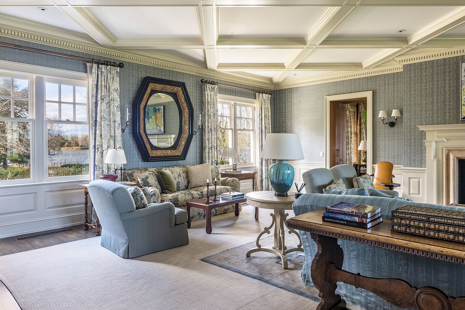This is a traditionally styled living room featuring blue patterned wallpaper, a coffered ceiling, and large windows that provide natural light. The room is furnished with comfortable seating, including sofas and armchairs upholstered in coordinating fabrics. A large, ornate mirror hangs on the wall, and a fireplace adds a cozy focal point.