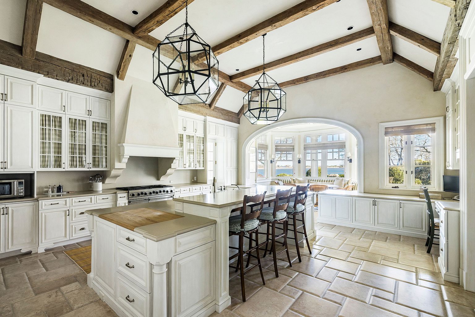 This is a spacious, high-end kitchen featuring custom white cabinetry, a large central island with bar seating, and exposed wooden beams on the vaulted ceiling. The kitchen is well-lit with natural light from large windows and stylish pendant lighting. The overall design exudes a luxurious and inviting atmosphere, perfect for both cooking and entertaining.