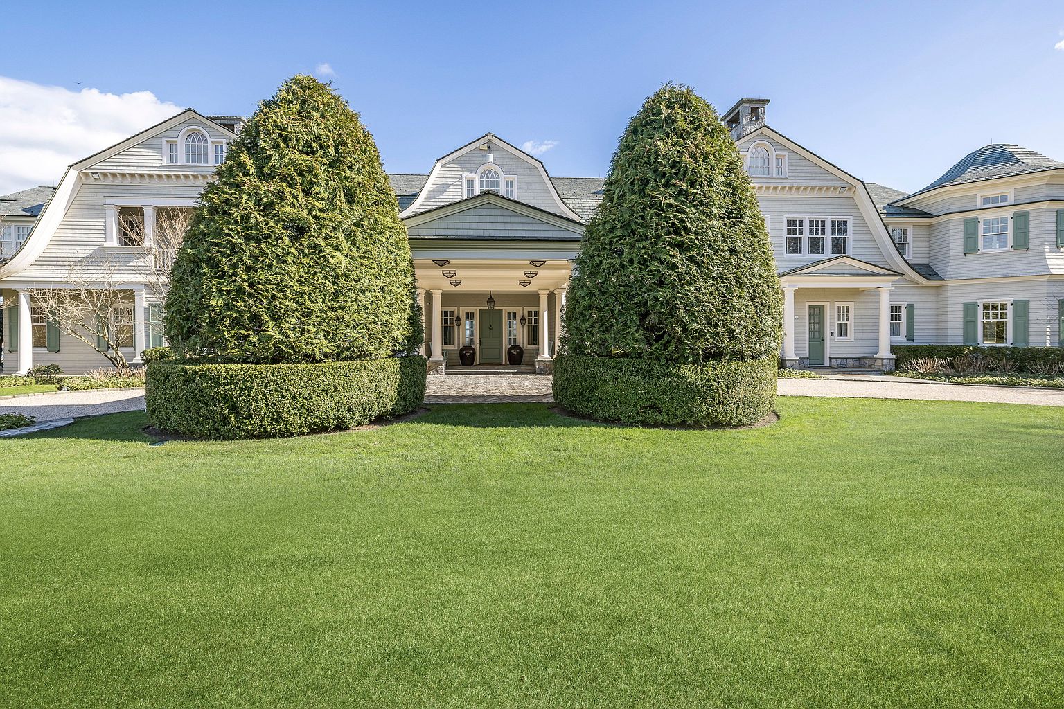This is a front view of a large, elegant house with a well-manicured lawn. The house features a light-colored exterior, multiple gabled roofs, and a covered entryway with columns. Two large, cone-shaped trees flank the entrance, adding to the property's curb appeal and creating a symmetrical composition.