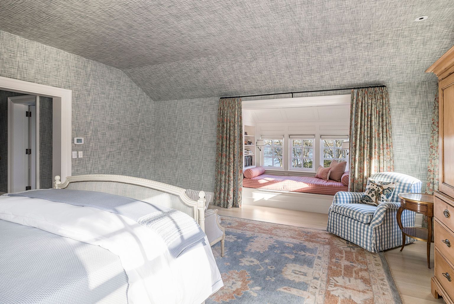 This is a primary bedroom featuring a serene and sophisticated design. The room includes a bed with a light-colored frame, a cozy window seat with floral curtains, and a comfortable armchair. The walls and ceiling are covered in a textured wallpaper, creating a cohesive and elegant aesthetic.