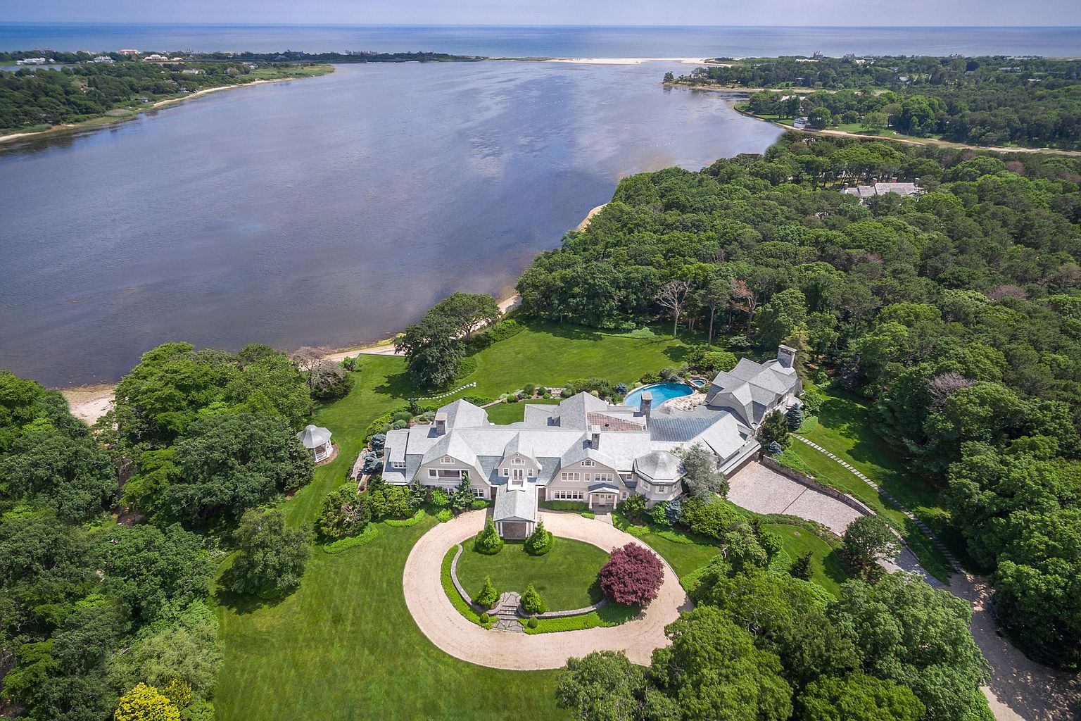 This aerial view showcases a grand estate situated on a lush, waterfront property. The sprawling residence features a complex roofline, a circular driveway, and meticulously manicured lawns. A swimming pool is nestled near the house, and the property is bordered by dense trees and a serene body of water, offering both privacy and stunning views.