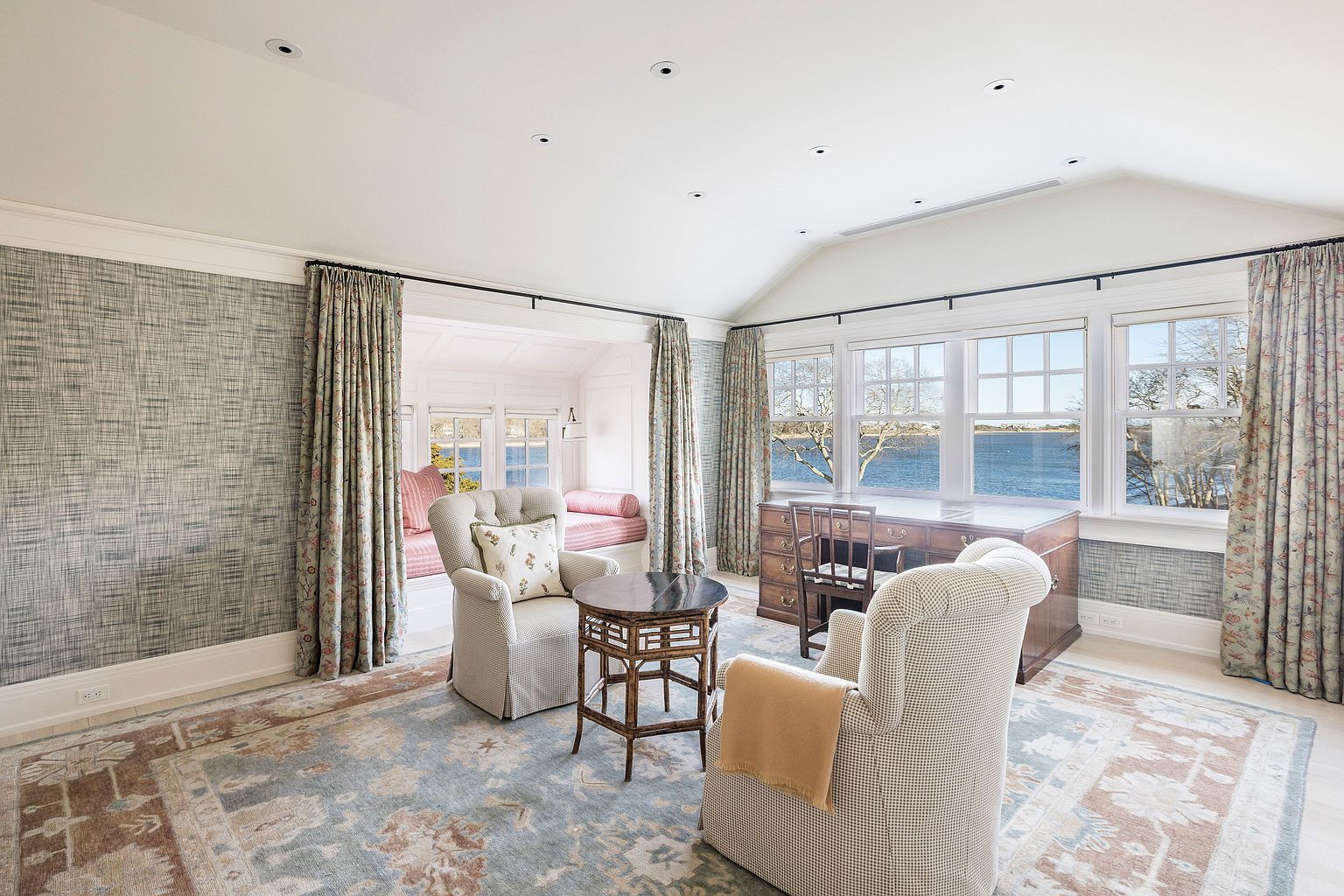 This is an interior shot of a well-lit office or study. The room features a built-in window seat, a desk with a view of the water, and two comfortable armchairs. The decor includes patterned wallpaper and curtains, creating a cozy and inviting atmosphere, complemented by a patterned rug that ties the room together.