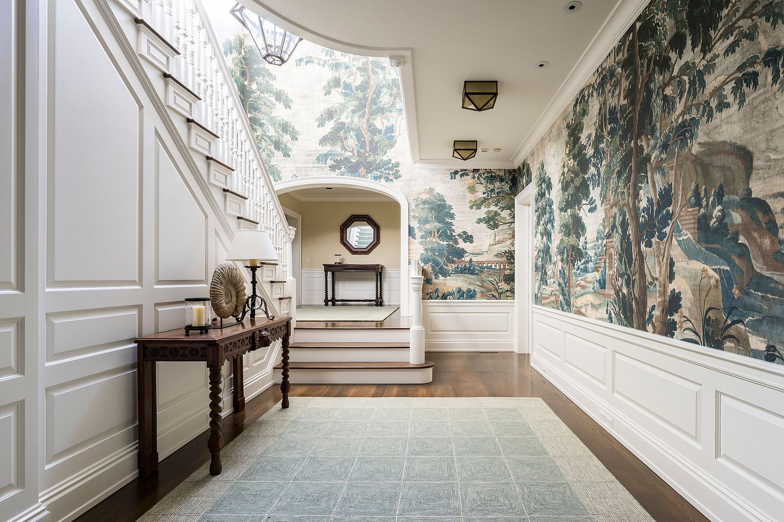 This interior shot showcases a grand hallway with a staircase, featuring elegant white wainscoting and a striking wallpaper mural depicting a landscape scene. A dark wood console table with a lamp and decorative items sits to the left, while a patterned rug adds texture to the hardwood floor. The space is well-lit and exudes a sophisticated, traditional style.