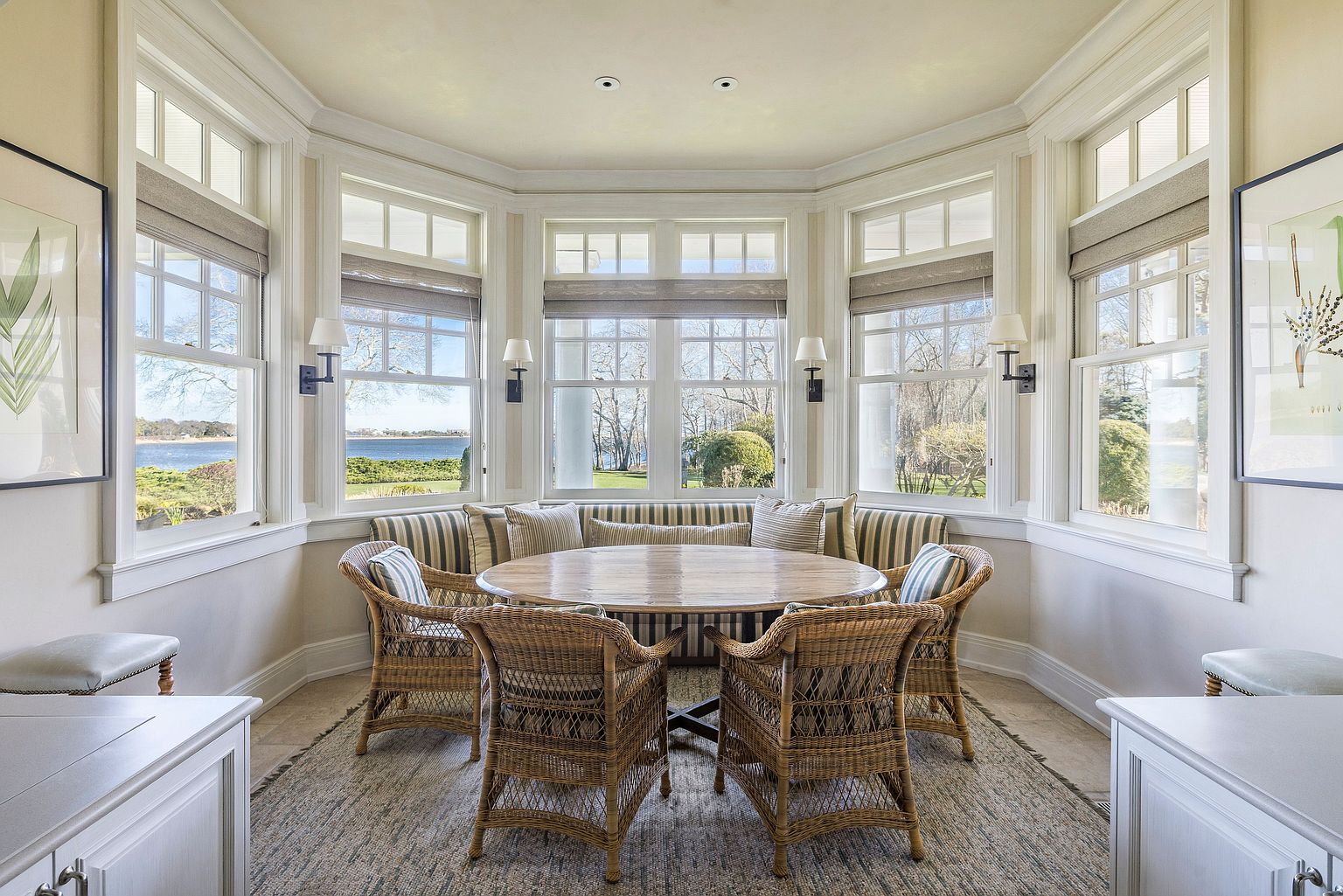 This sunroom dining area features a round wooden table surrounded by wicker chairs and a built-in banquette seating. Large windows offer panoramic views of the water and landscape, complemented by neutral-toned roman shades and sconce lighting. The space is bright and airy, creating an inviting atmosphere for dining.
