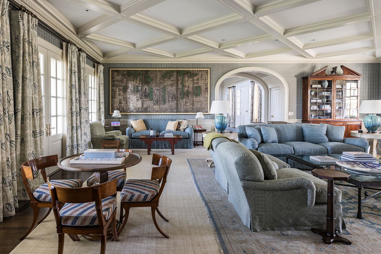 This is a spacious and elegant living room featuring a coffered ceiling, patterned wallpaper, and multiple seating areas. The room is furnished with comfortable sofas, armchairs, and a round table with chairs, creating a welcoming and sophisticated atmosphere. A large piece of artwork hangs above one of the sofas, adding a focal point to the room.