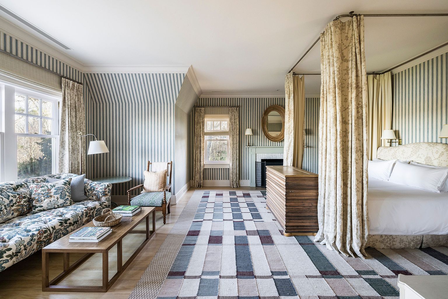 This is a spacious primary bedroom featuring striped wallpaper and a four-poster bed with elegant curtains. The room includes a seating area with a floral sofa and a wooden coffee table, as well as a fireplace with a decorative mirror above it. A patterned rug adds texture to the hardwood floor, creating a cozy and sophisticated atmosphere.