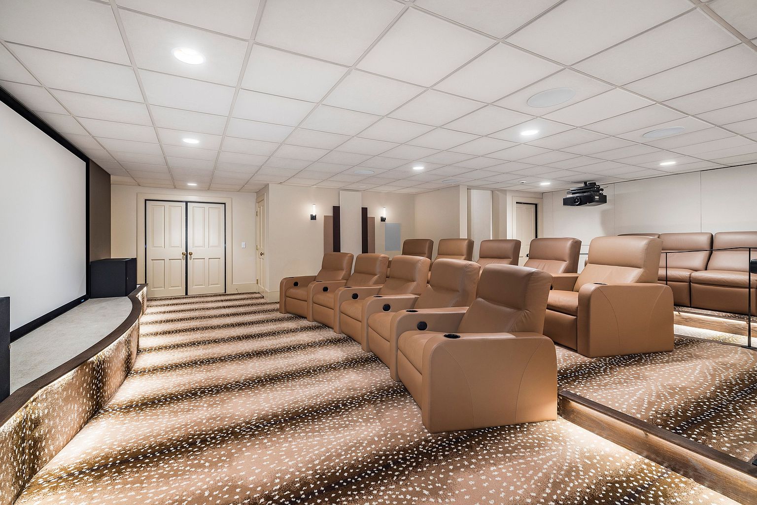 This is an interior shot of a home theater, featuring rows of tan leather reclining chairs with cup holders, arranged on tiered platforms covered with a patterned carpet. A large screen is visible on the left, and a projector is mounted on the ceiling. The room has a drop ceiling with recessed lighting, creating a comfortable and immersive viewing experience.