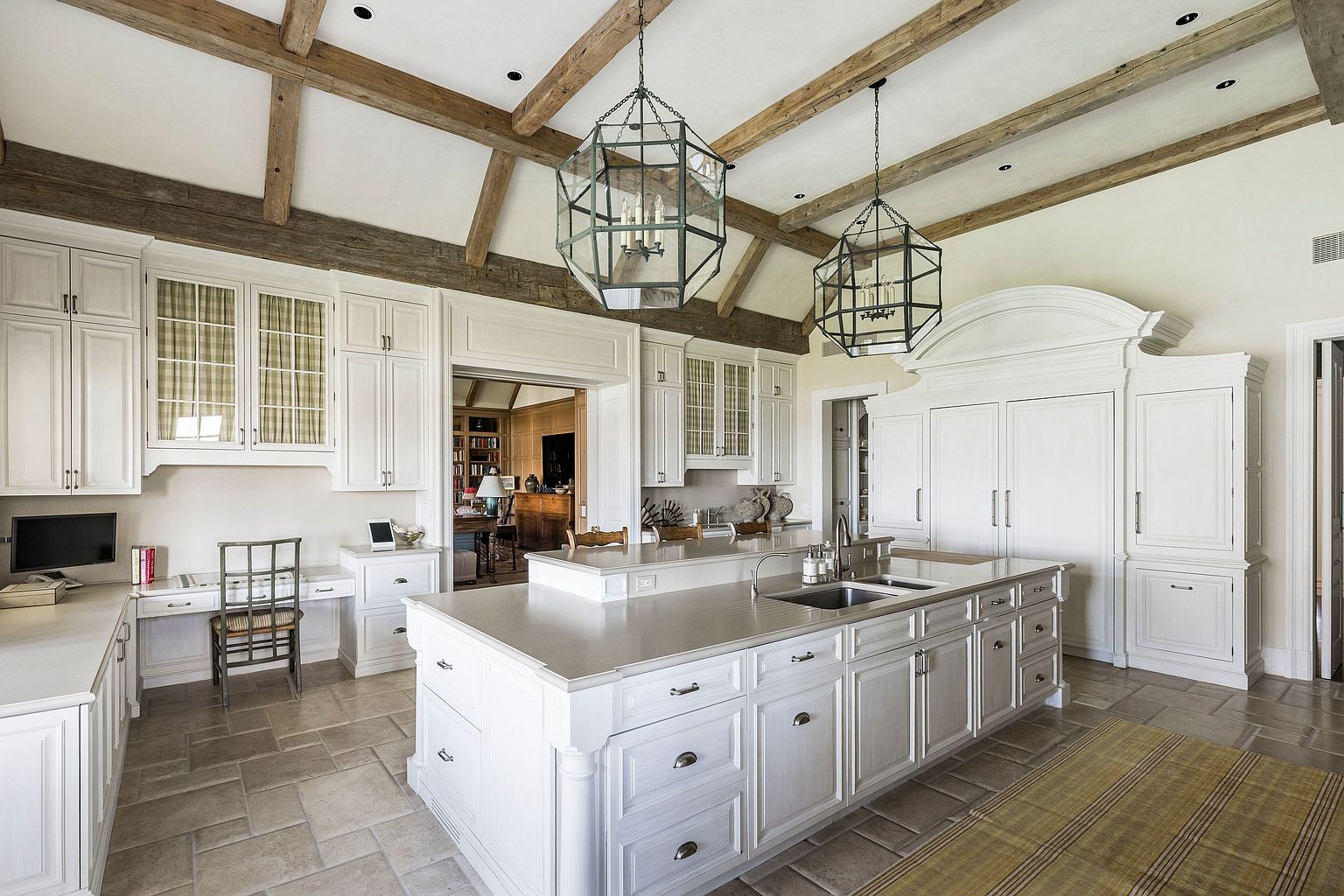 This is a spacious kitchen featuring white cabinetry, a large central island with a sink, and two decorative pendant lights. The kitchen also includes a built-in desk area and wooden ceiling beams, adding a touch of rustic charm to the elegant design. The overall impression is one of a well-appointed and functional space.
