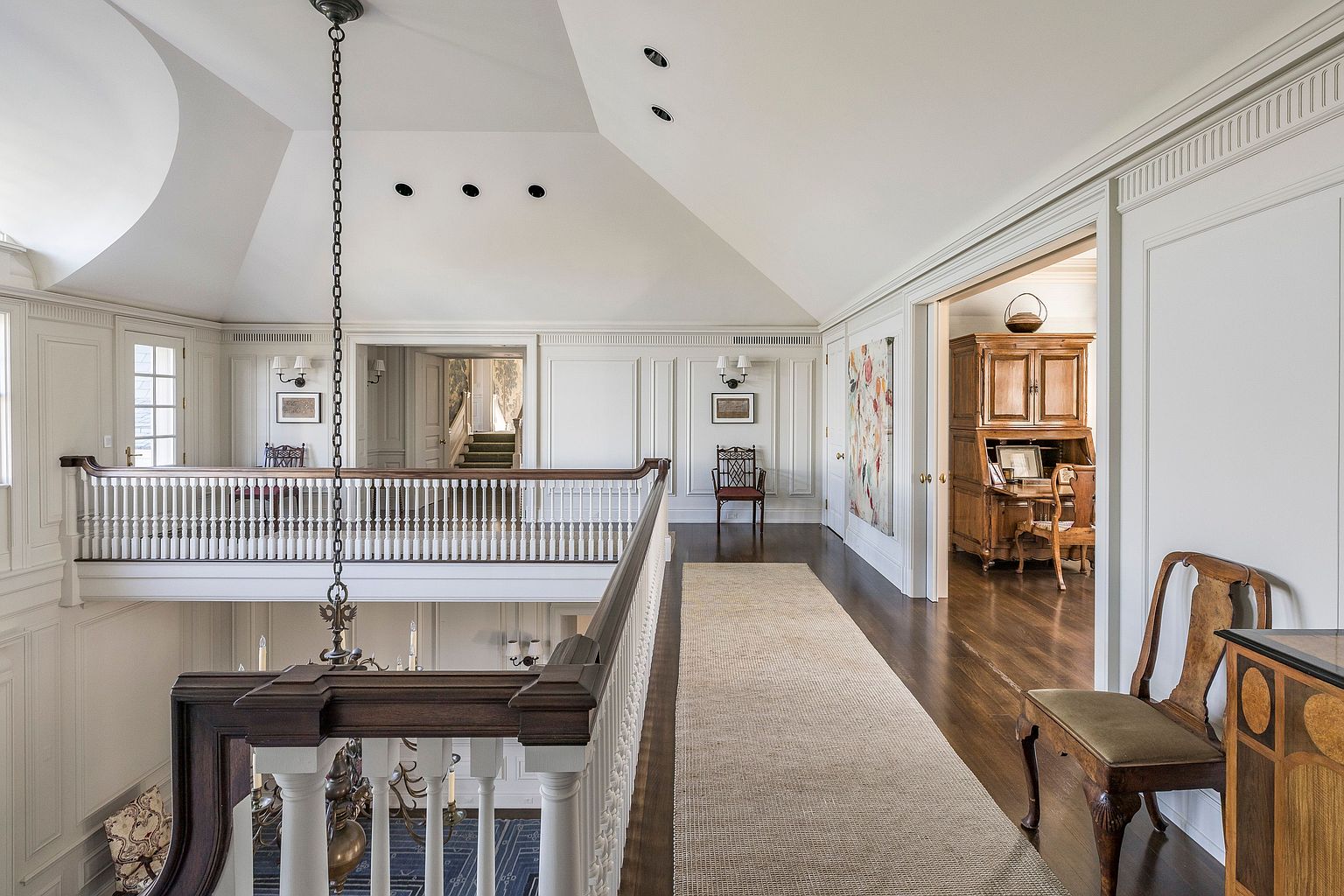 This interior shot showcases a grand hallway with a staircase and a second-floor landing. The hallway features white paneled walls, dark hardwood floors, and a light-colored runner rug. A chandelier hangs from the ceiling, and a glimpse into an adjacent room reveals a wooden desk and cabinet, suggesting a home office or study.