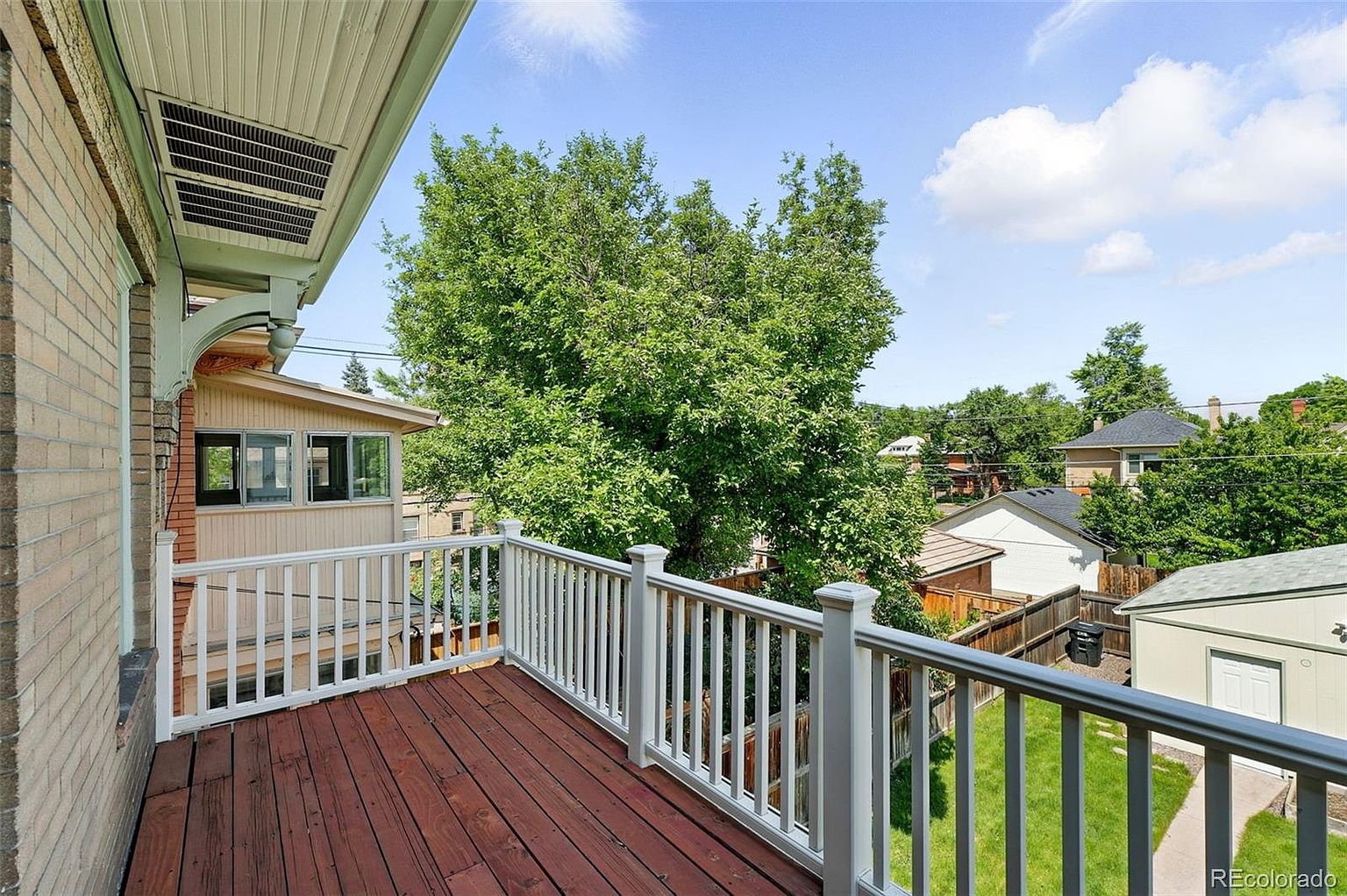 This image showcases a charming balcony with a wooden deck and white railing, offering a pleasant outdoor space. The view extends to a lush green yard and neighboring houses, framed by a large, leafy tree. The balcony appears to be attached to a brick building, adding to its character and appeal.