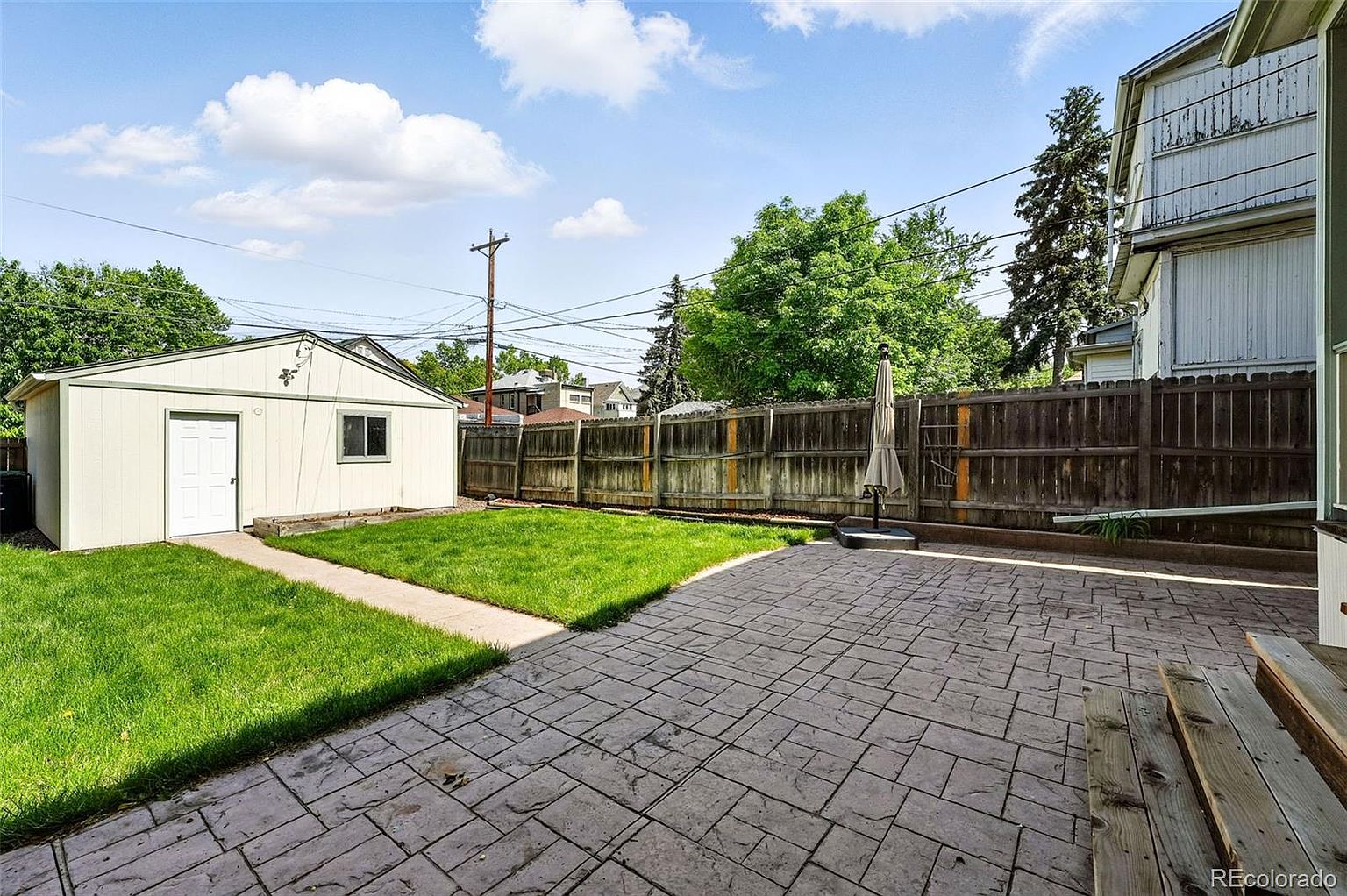 This image showcases a well-maintained backyard featuring a combination of elements. A concrete patio provides a functional outdoor space, complemented by a grassy area adjacent to a small outbuilding or shed. A wooden fence encloses the yard, offering privacy, while mature trees and a clear sky create a pleasant atmosphere.