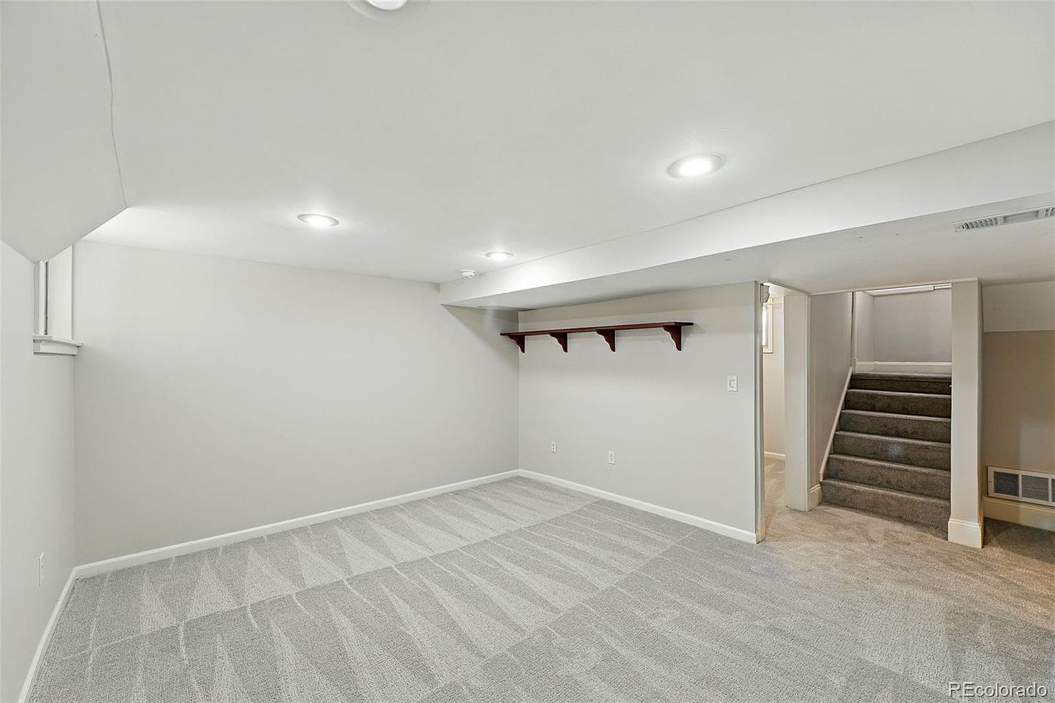 The image showcases a finished basement area with neutral gray carpeting and light gray walls. A set of carpeted stairs leads to another level, and a dark wood shelf is mounted on the wall. Recessed lighting provides ample illumination, creating a clean and inviting space.