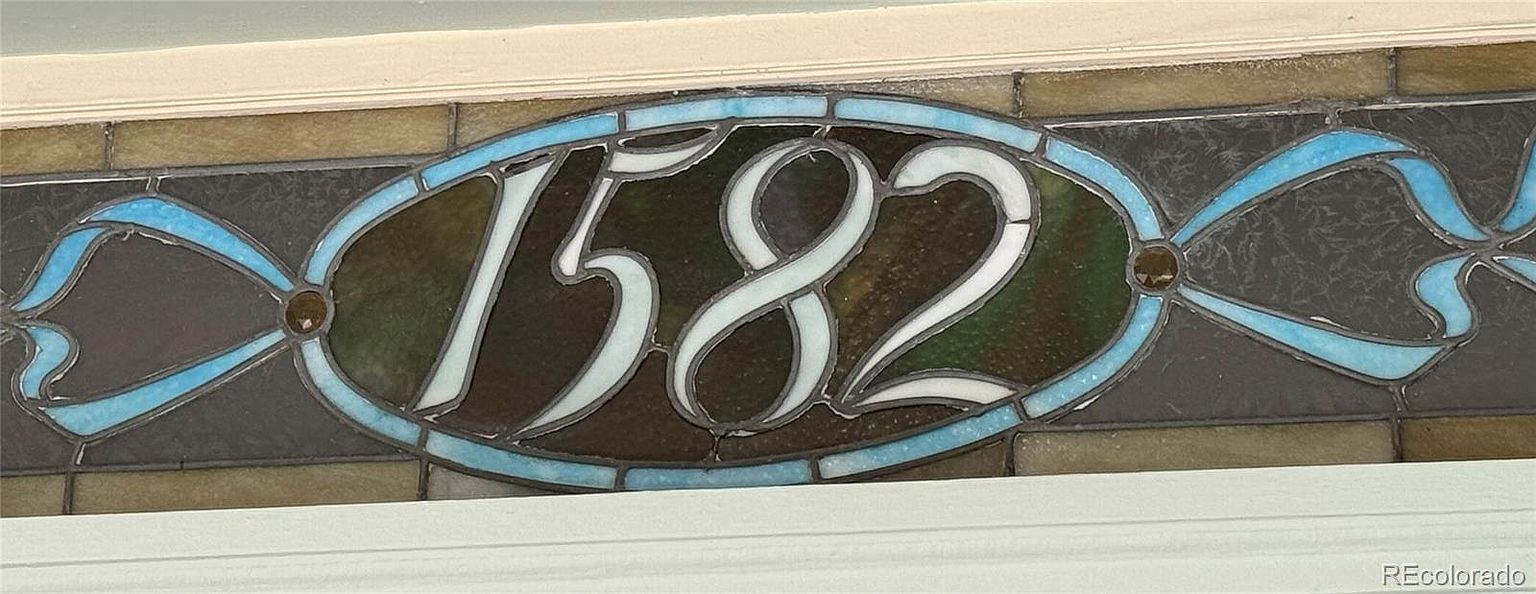 The image showcases a stained glass window displaying the house number "1582". The window is oval-shaped with a blue ribbon design around the perimeter, adding a touch of elegance and vintage charm. The numbers are in a clear, contrasting color, making them easily visible and contributing to the property's curb appeal.