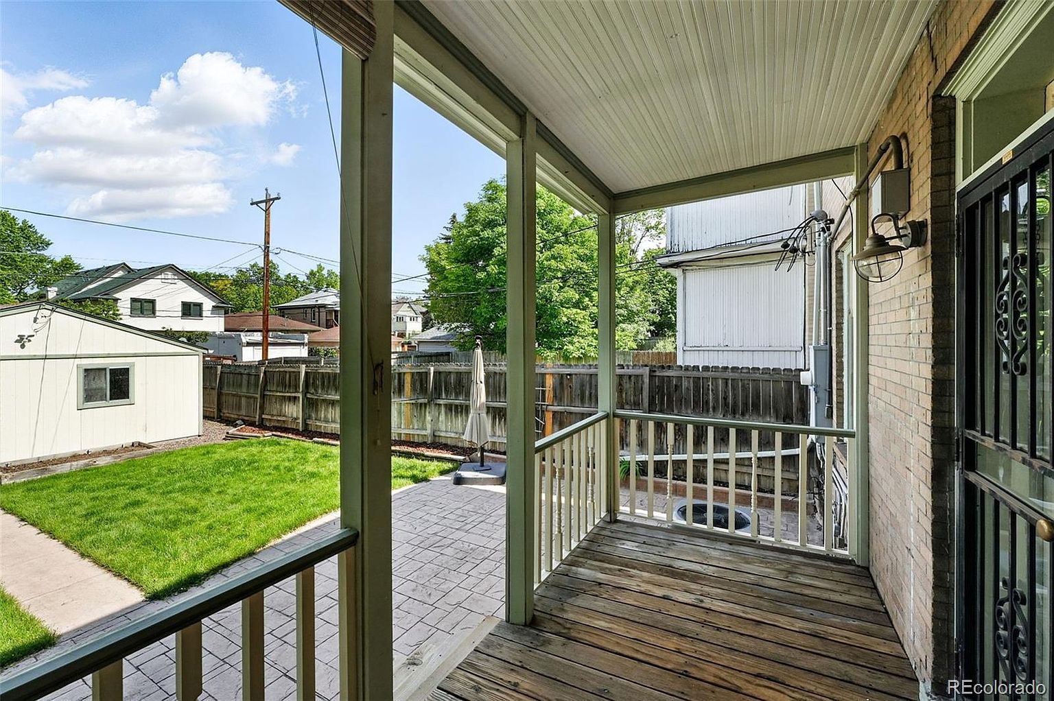 This image showcases a charming outdoor patio/deck area. The deck features wooden flooring and a railing, leading to a stone-paved patio and a well-maintained lawn. The view extends to a fenced backyard with a shed and neighboring houses, creating a sense of privacy and suburban comfort.