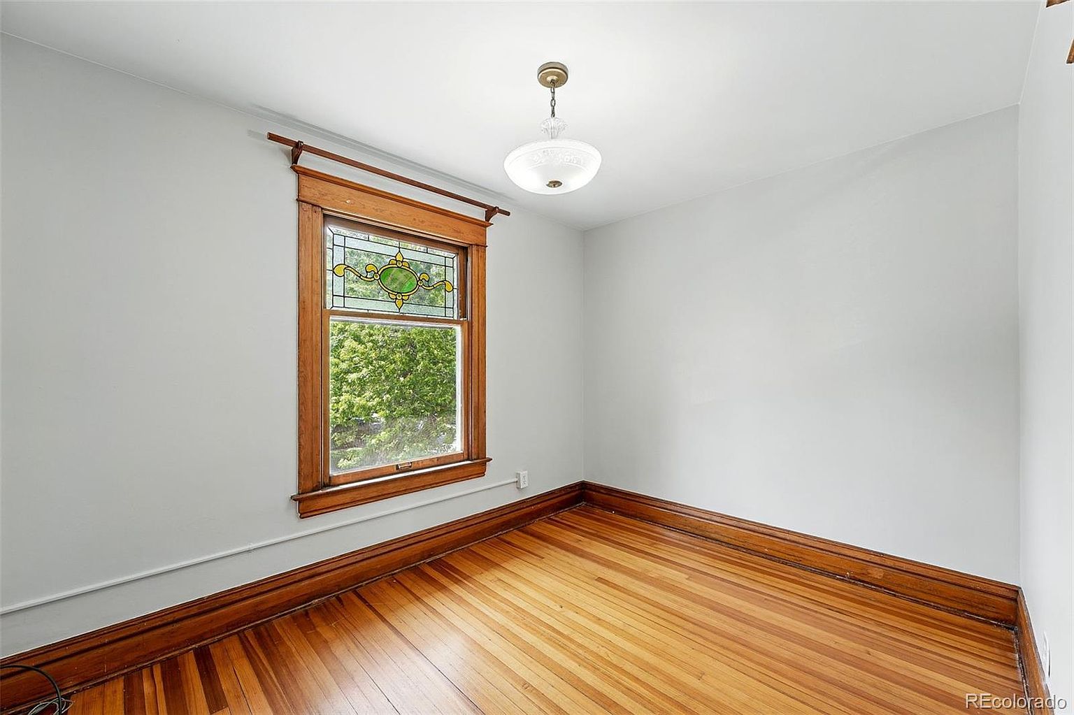This is an interior shot of a bedroom featuring hardwood floors and a stained glass window. The walls are painted a neutral gray, and a vintage-style light fixture hangs from the ceiling. The room appears bright and airy, with a classic, understated charm.