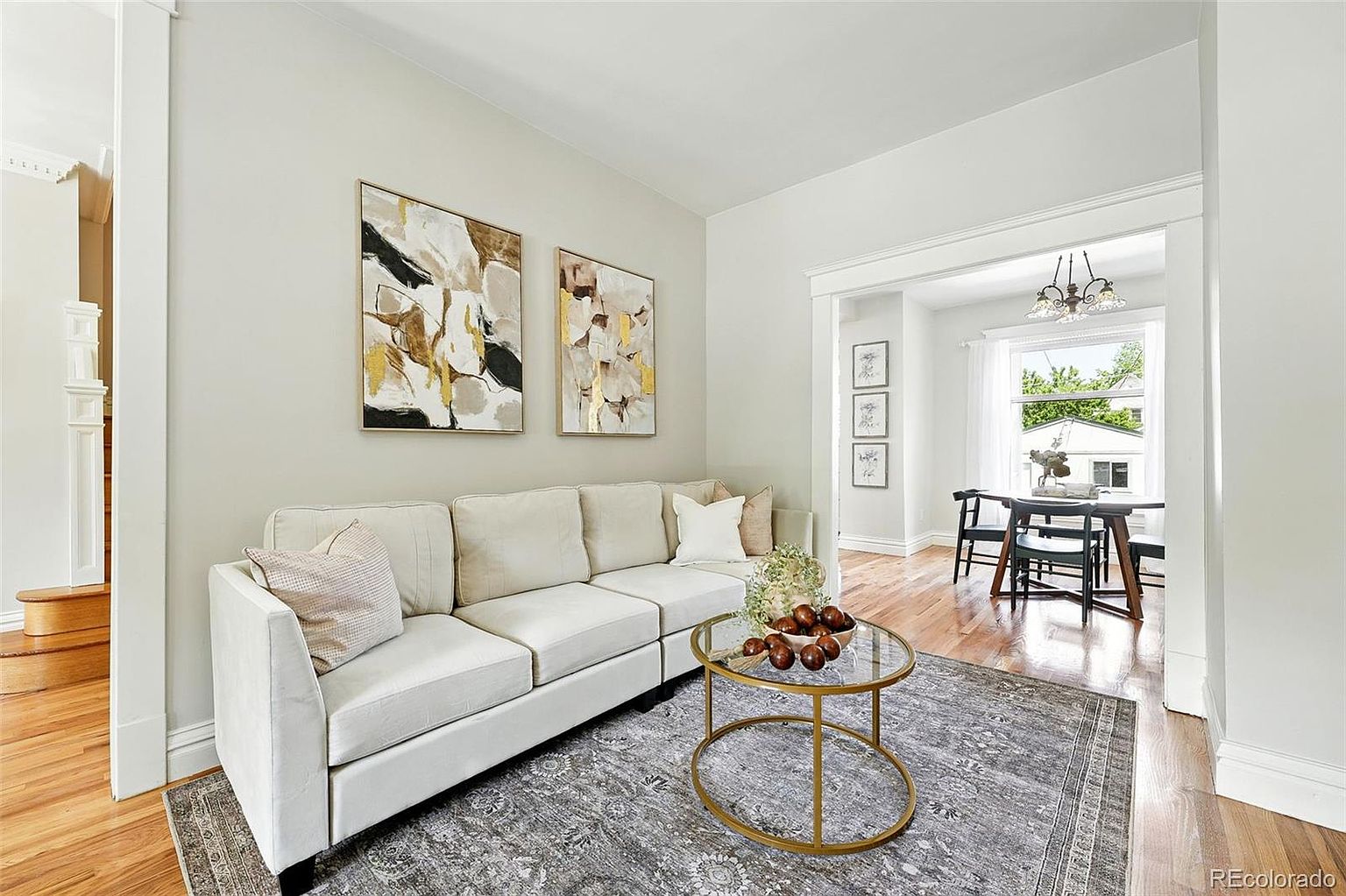 This is an interior shot of a living room featuring a cream-colored sofa, a round glass coffee table with a gold frame, and a gray patterned rug. Two abstract paintings hang on the wall above the sofa. The room opens into a dining area, creating an open-concept living space with hardwood floors throughout.