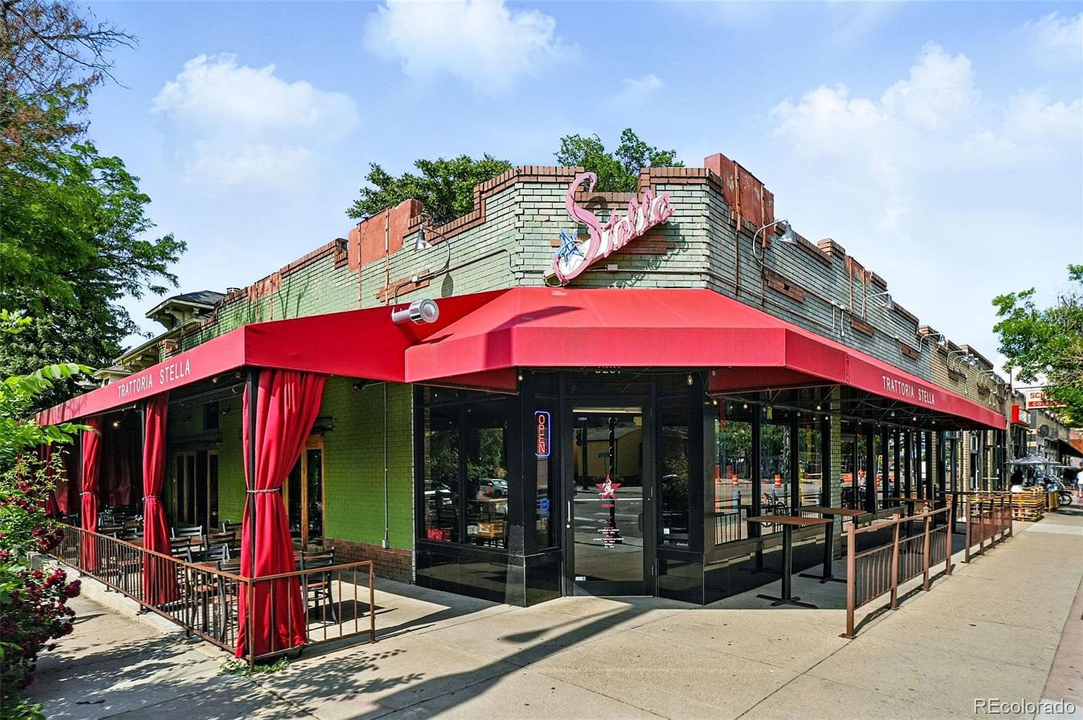 The image showcases the front exterior of Trattoria Stella, a restaurant with a distinctive architectural style. The building features a red awning that extends over the entrance and outdoor seating area, complemented by red curtains. The facade is a mix of green and brick, with a neon flamingo sign adding a playful touch. The overall impression is inviting and suggests a vibrant dining experience.