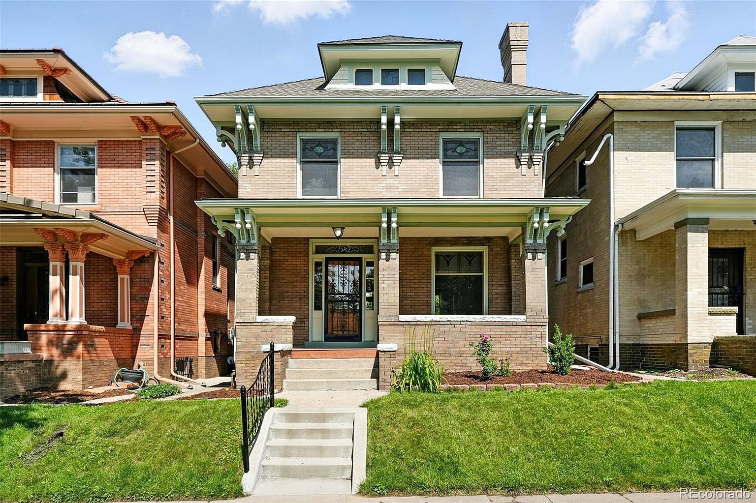 This is a front view of a two-story brick house with a covered porch. The house features a symmetrical facade with windows on both floors and decorative architectural details. The front yard is well-maintained with green grass and a concrete staircase leading to the porch, creating a welcoming and classic curb appeal.