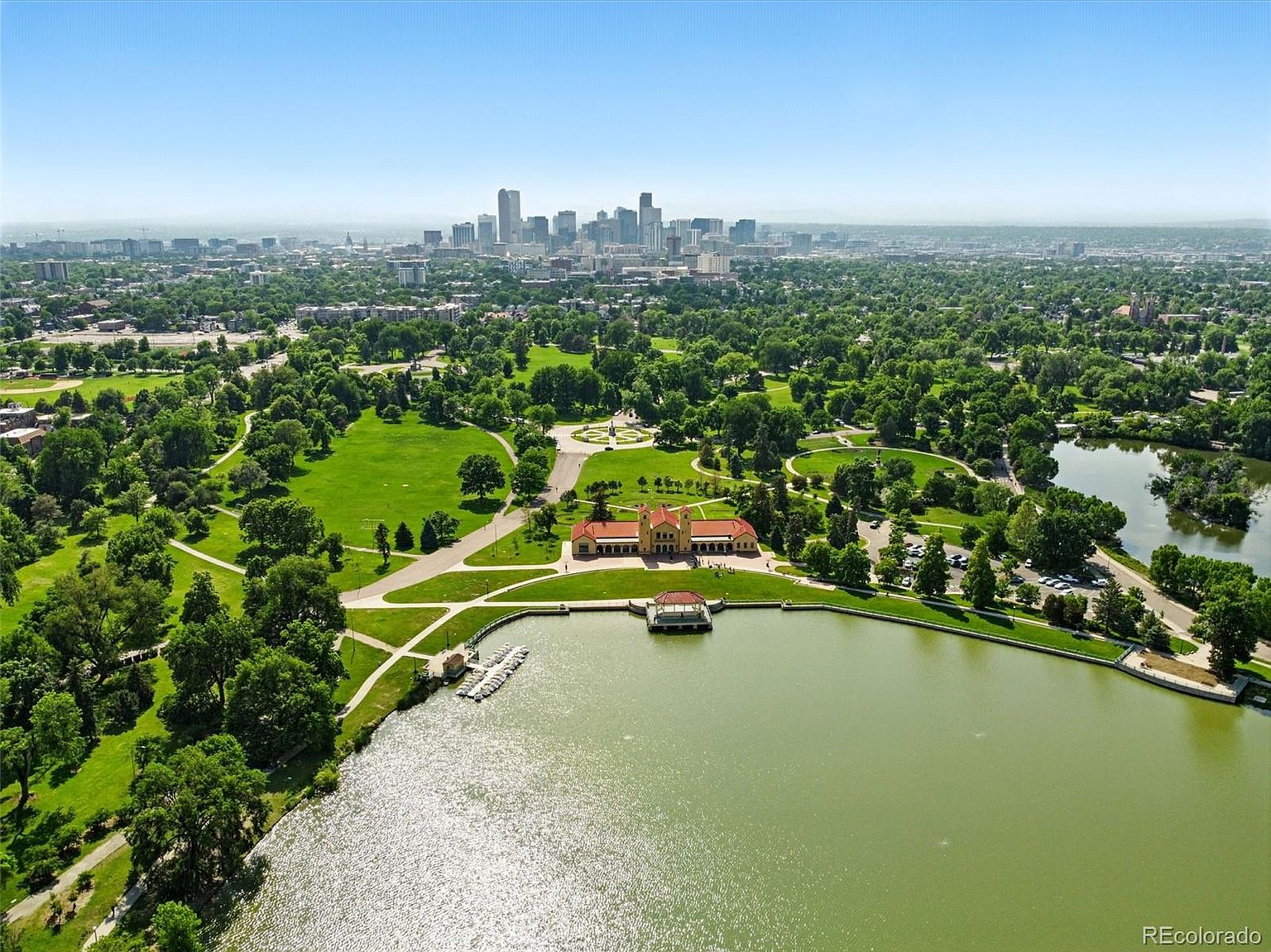 This aerial view showcases a large park with lush green spaces, mature trees, and a serene lake. A prominent building with a red-tiled roof sits near the water's edge, adding architectural interest. In the distance, the city skyline provides a stunning backdrop, emphasizing the park's proximity to urban amenities.