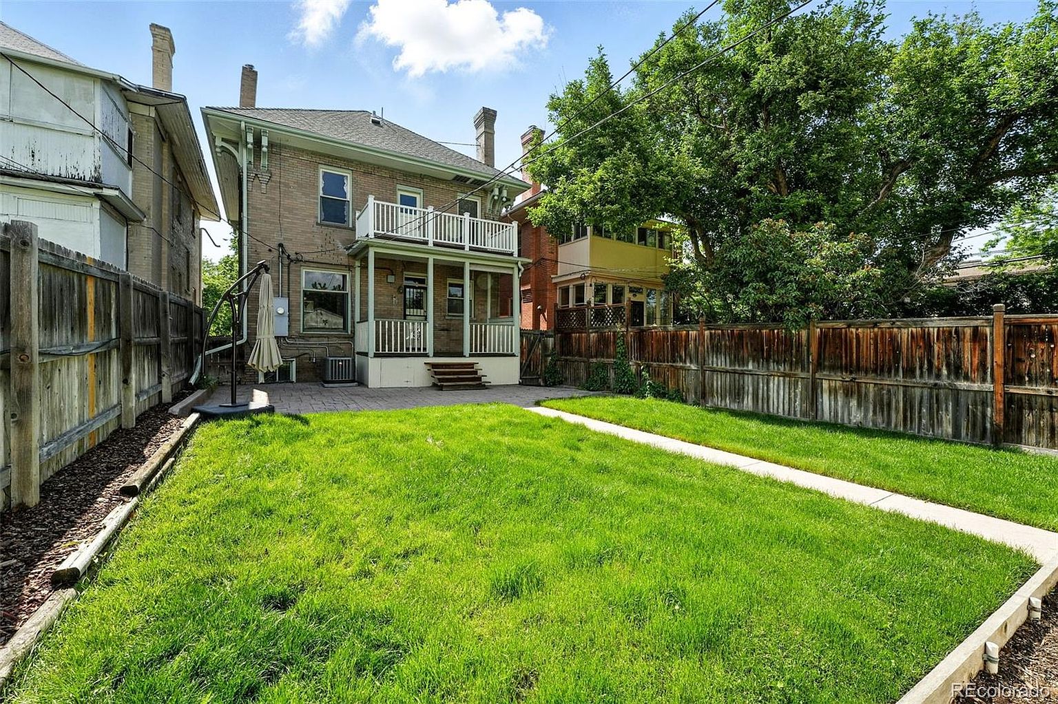 This image showcases the backyard of a two-story brick house, featuring a well-maintained lawn and a paved patio area. A wooden fence encloses the yard, providing privacy, while a pathway leads through the grass. The presence of mature trees adds to the property's appeal, creating a serene outdoor space.