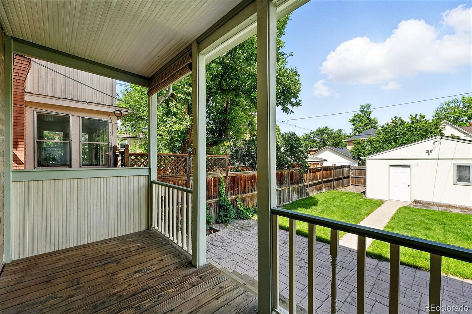 This image showcases a charming outdoor patio or deck area, featuring a wooden floor and railing. The view extends to a well-maintained backyard with a grassy lawn, a paved walkway, and a detached garage. The space offers a relaxing outdoor living area with a view of the backyard.
