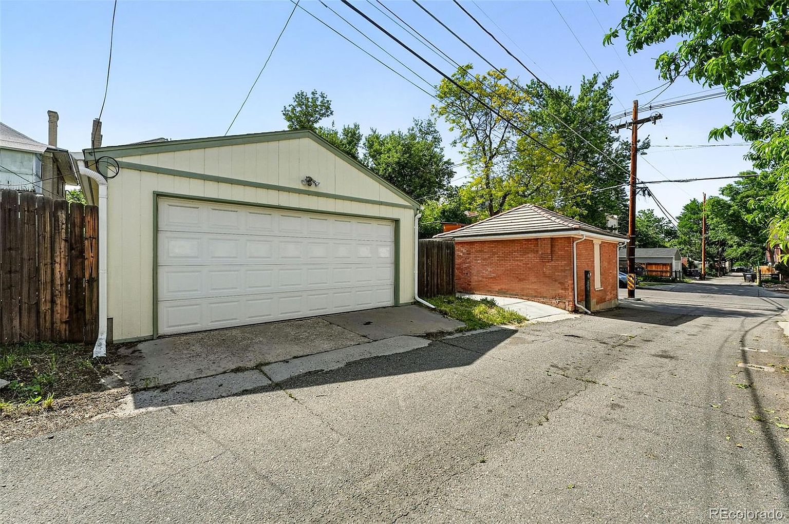 The image showcases a detached garage and a small brick outbuilding, likely a shed or additional storage space, situated along a narrow alleyway. The garage features a white door with a paneled design and light-colored siding, while the brick structure has a contrasting tile roof. The alleyway is paved and shows signs of wear, with utility poles and trees visible in the background, creating a functional yet somewhat aged aesthetic.