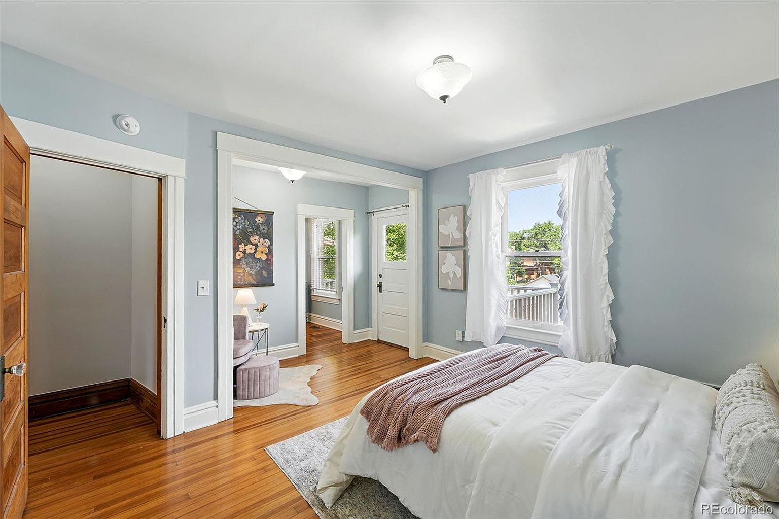 This is a bright and airy primary bedroom featuring light blue walls, hardwood floors, and white trim. The room includes a bed with white linens and a pink throw, two windows with white ruffled curtains, and an adjacent sitting area with a floral wall hanging. The overall impression is clean, comfortable, and inviting.