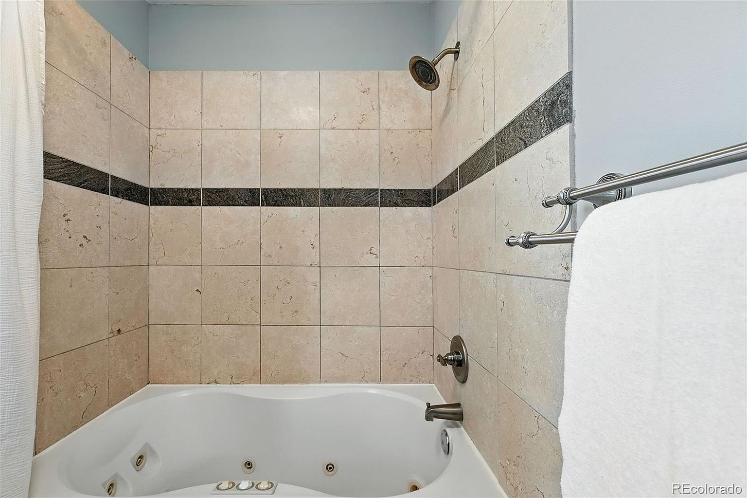 This is a well-lit primary bathroom featuring a jacuzzi tub with tile surround and a shower. The tiles are a light beige color with a dark accent stripe. A towel rack with a white towel is visible on the right side of the image.