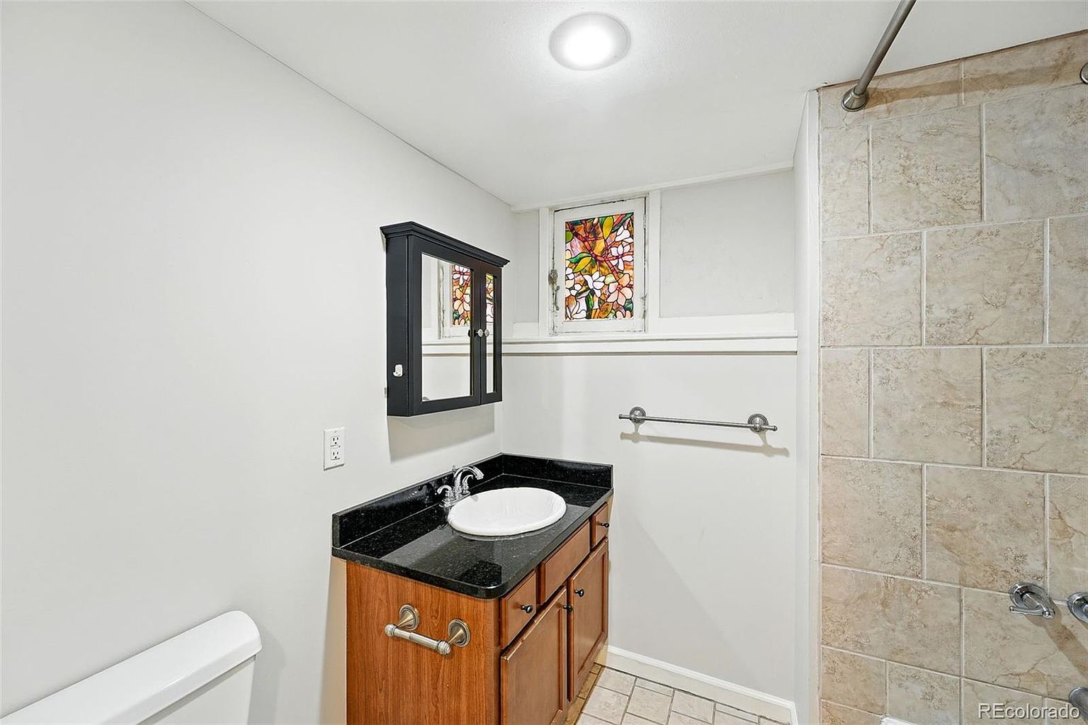 This is a bathroom featuring a vanity with a dark countertop and wooden cabinets, a white sink, and a black framed medicine cabinet. A stained glass window adds a unique touch, and the shower area is tiled. The walls are painted a neutral color, creating a clean and functional space.
