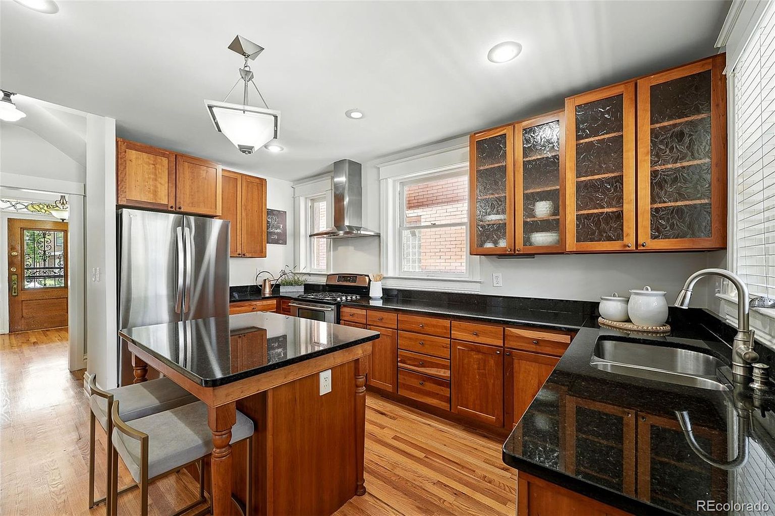 This is a well-lit kitchen featuring wooden cabinetry with black countertops and stainless steel appliances. A kitchen island with bar seating is centrally located, and the hardwood flooring adds warmth to the space. The kitchen has a classic and functional design, suitable for both cooking and entertaining.