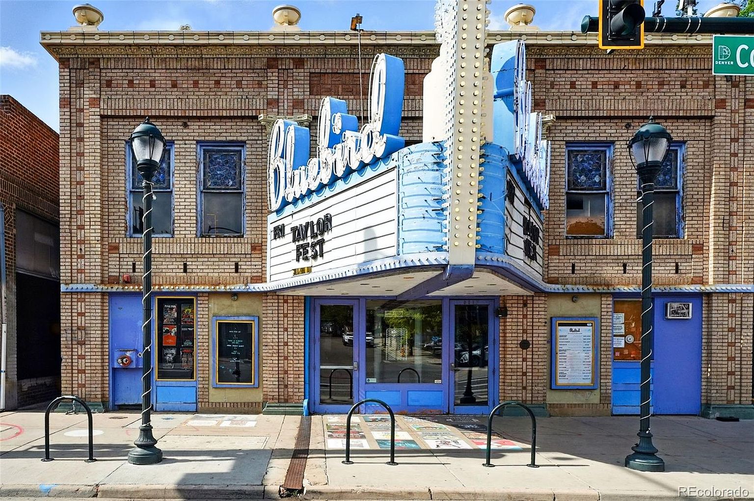 This is a front view of the Bluebird Theater, a brick building with a distinctive blue and white marquee. The facade features classic architectural details, including decorative brickwork and vintage-style lampposts. The entrance is framed by blue doors and windows, creating an inviting and unique property.